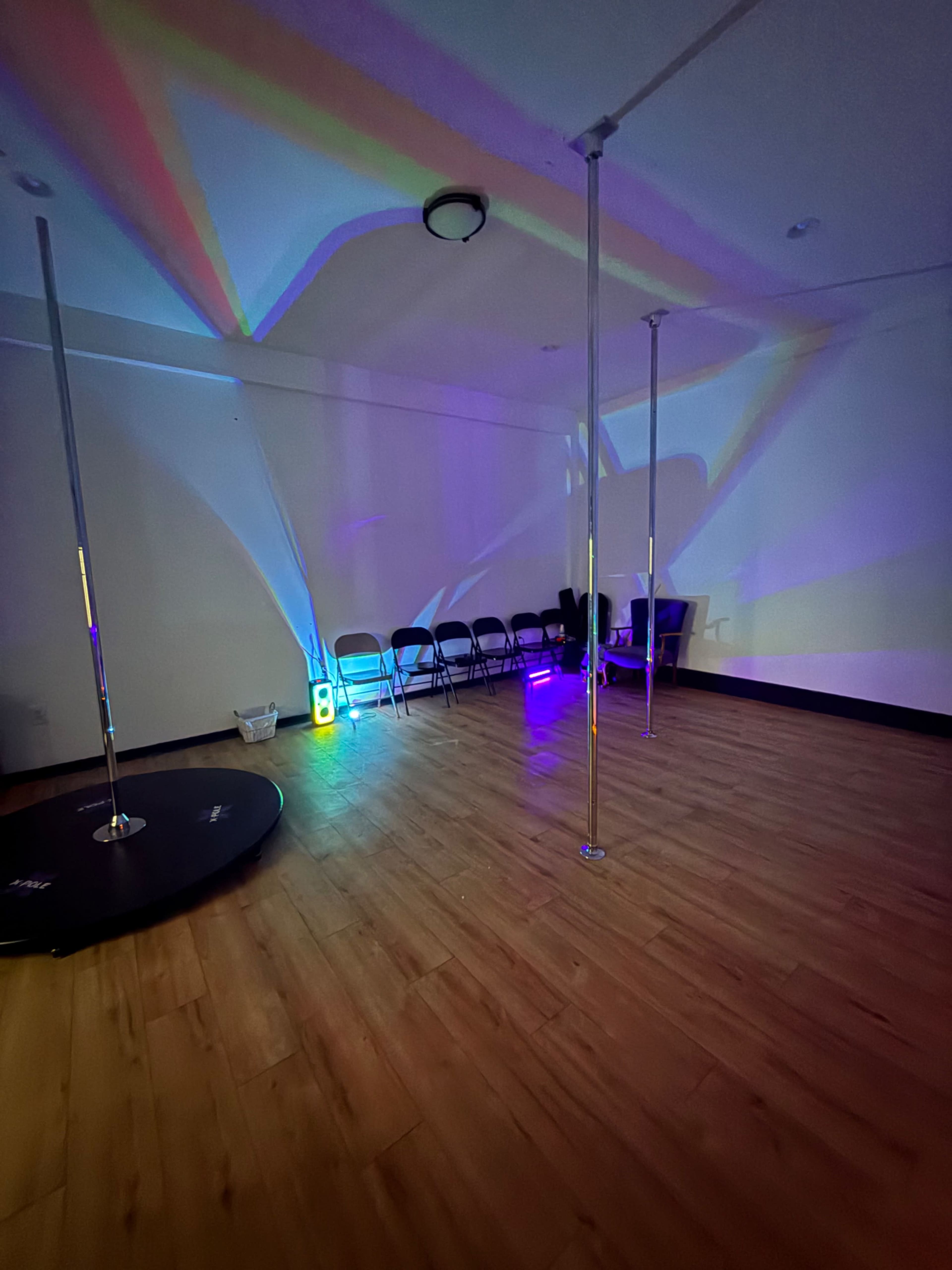 The image shows an indoor space with a wooden floor, a pole in the center, several chairs lined up against the wall, and colorful light projections casting shadows on the walls.