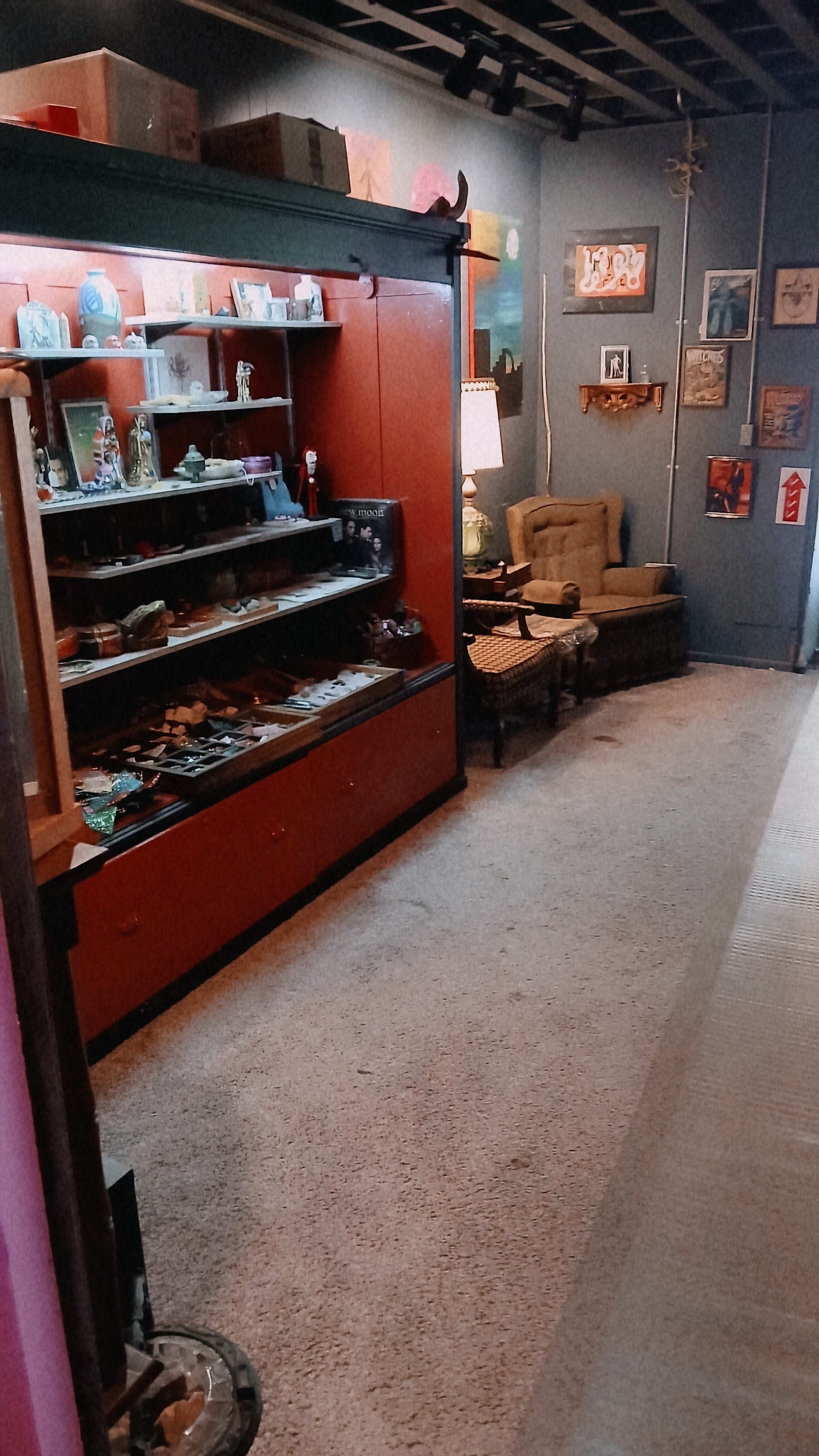 The image shows a room with a display case filled with various items on one side and a seating area featuring an armchair and lamp on the opposite side.