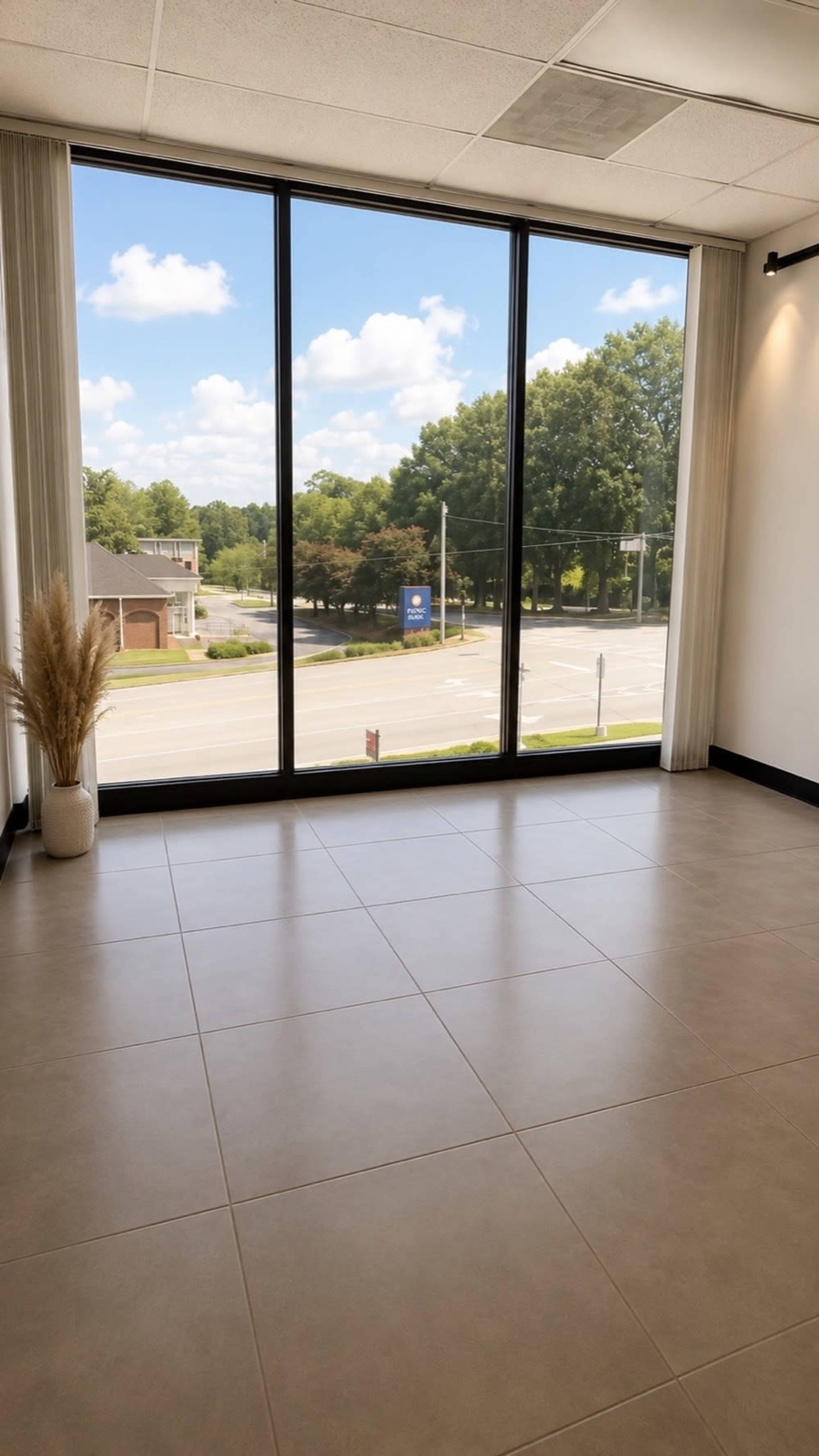 The image shows a spacious room with large windows offering a view of a tree-lined street under a blue sky.