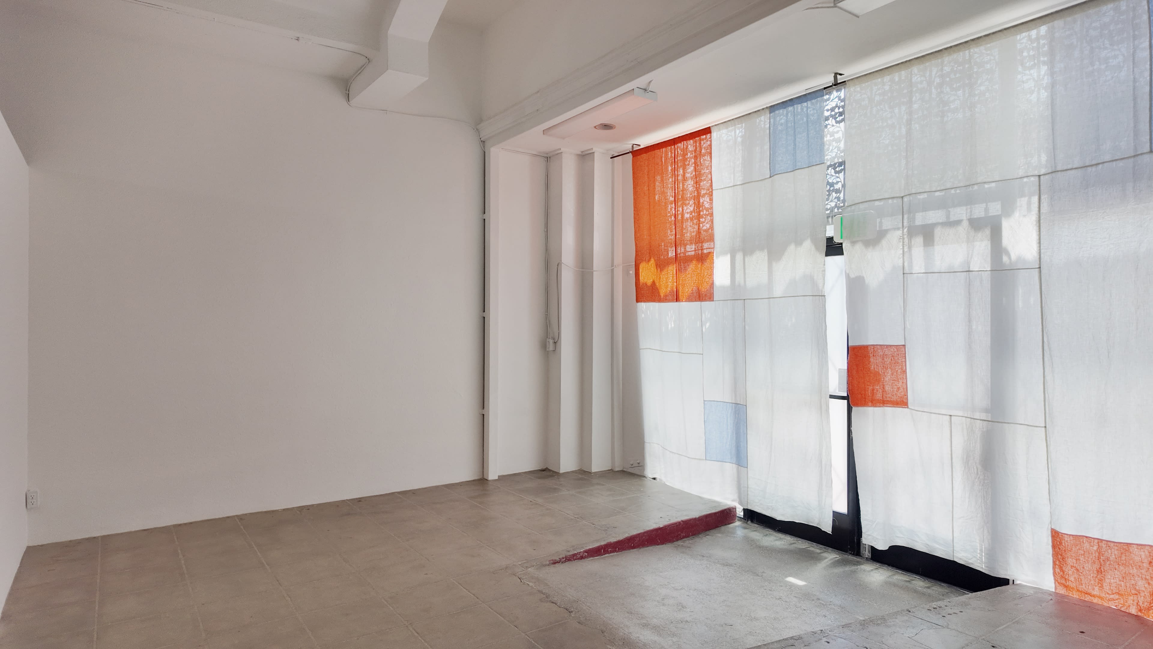 A sparsely furnished room features a large window covered by colorful sheer curtains, filtering light into the space.