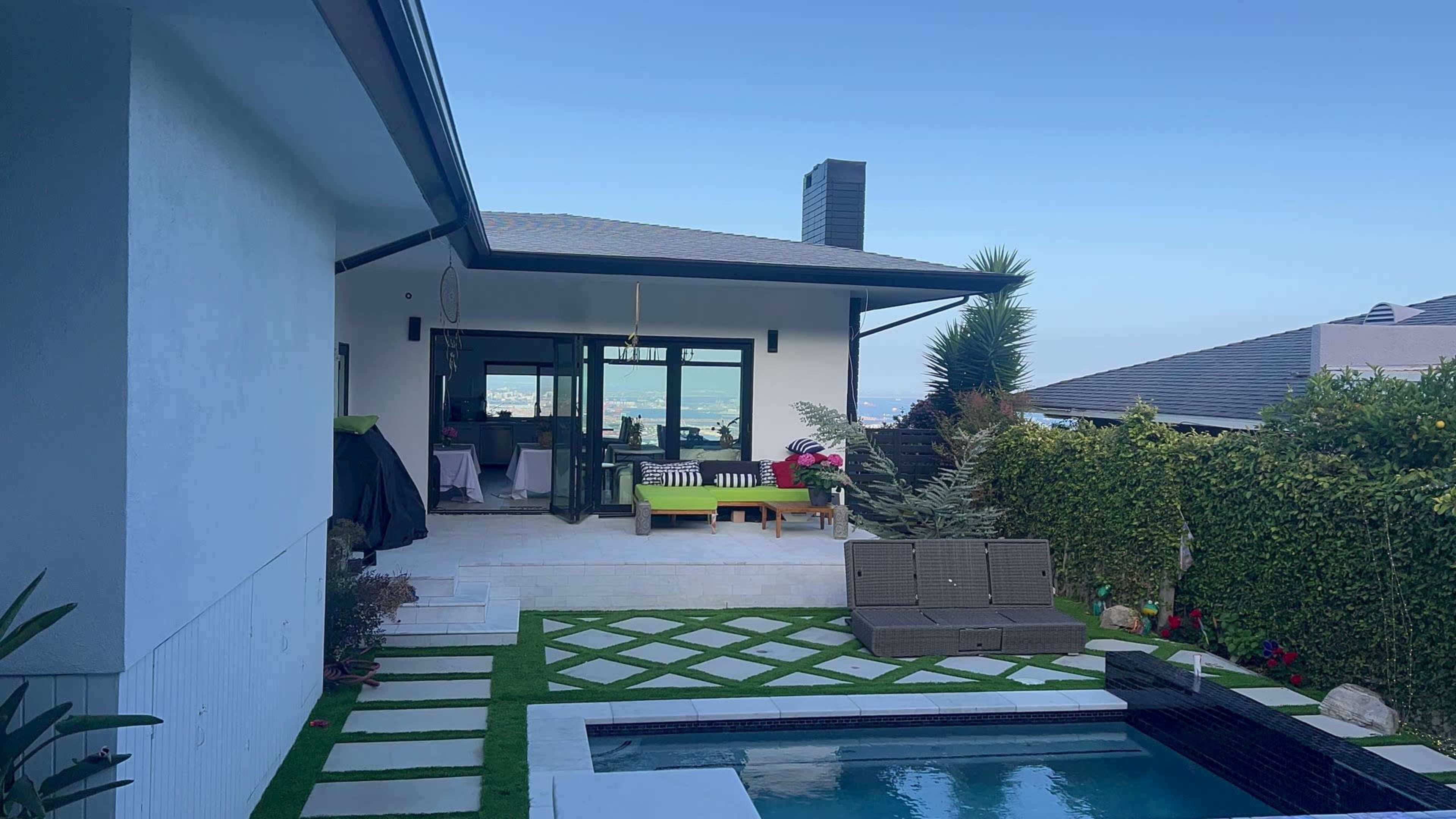 The image shows a modern backyard with a swimming pool, green lawn, and patio furniture, framed by a white house and views of a distant body of water.