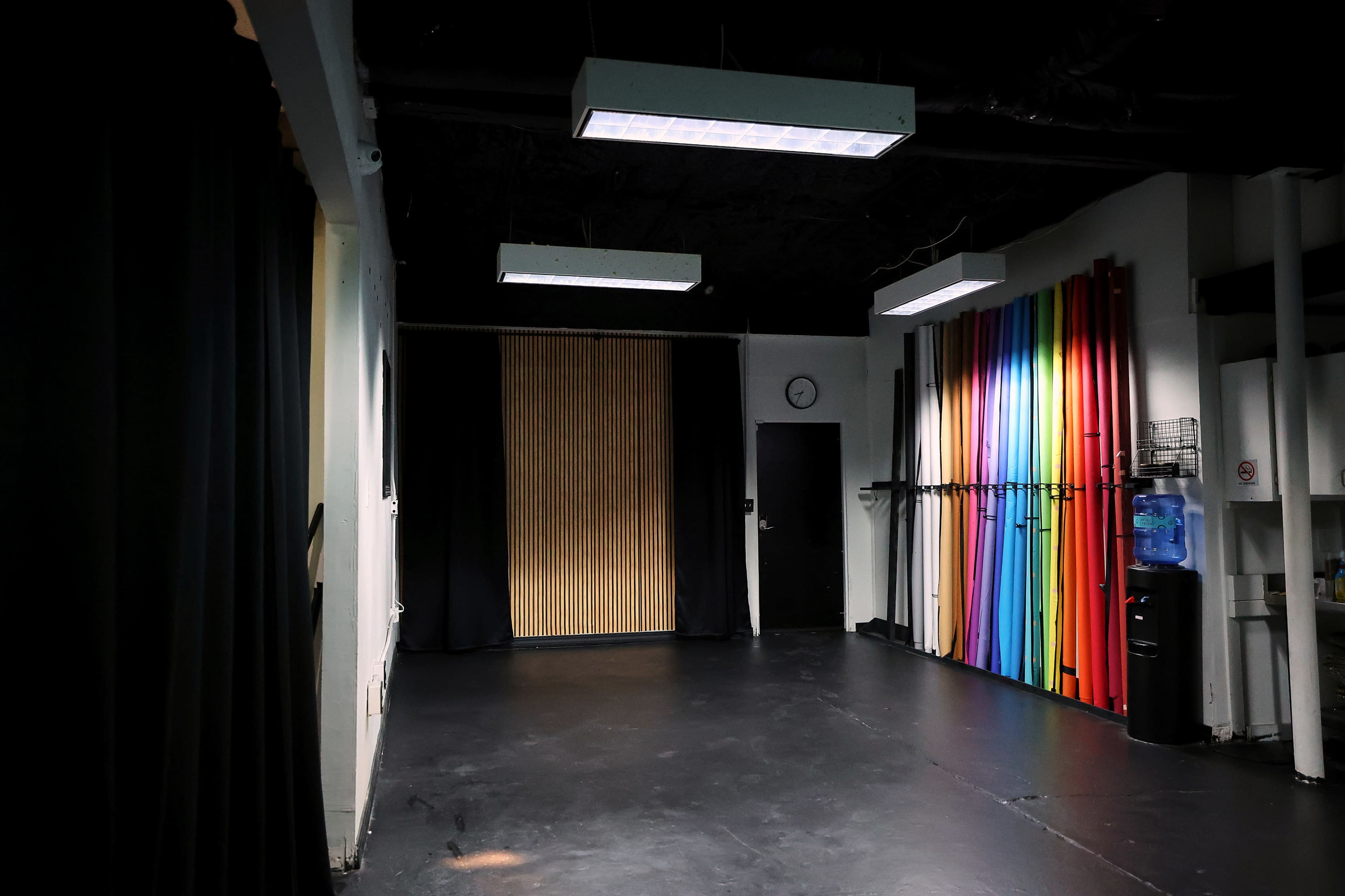 The image shows a spacious, dimly lit studio with a wooden panel in the center, surrounded by brightly colored vertical rolls of material on one side.