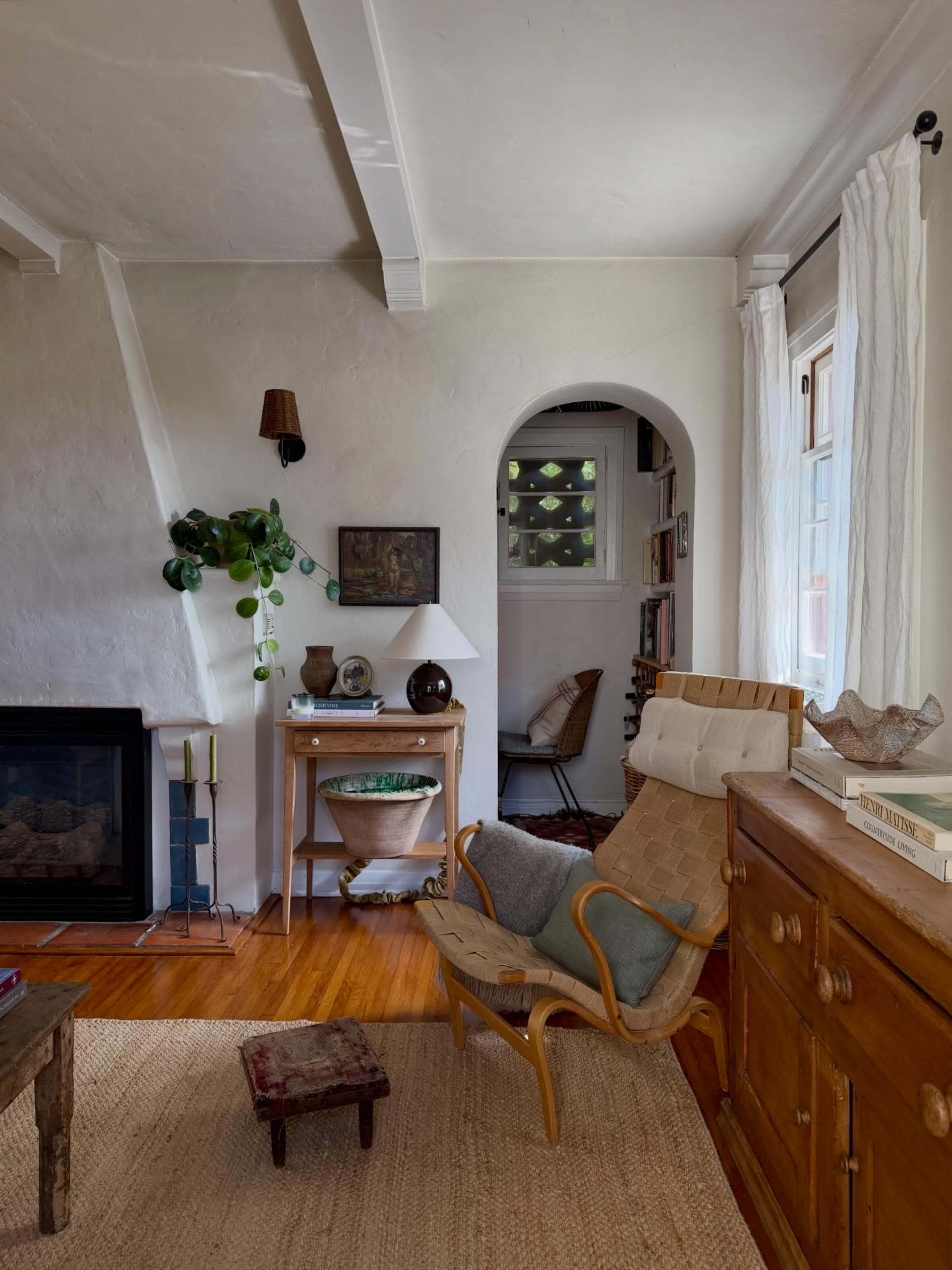 The image shows a cozy interior of a living room featuring a comfortable chair, a wooden table, and a fireplace, with plants and decorative items on display.