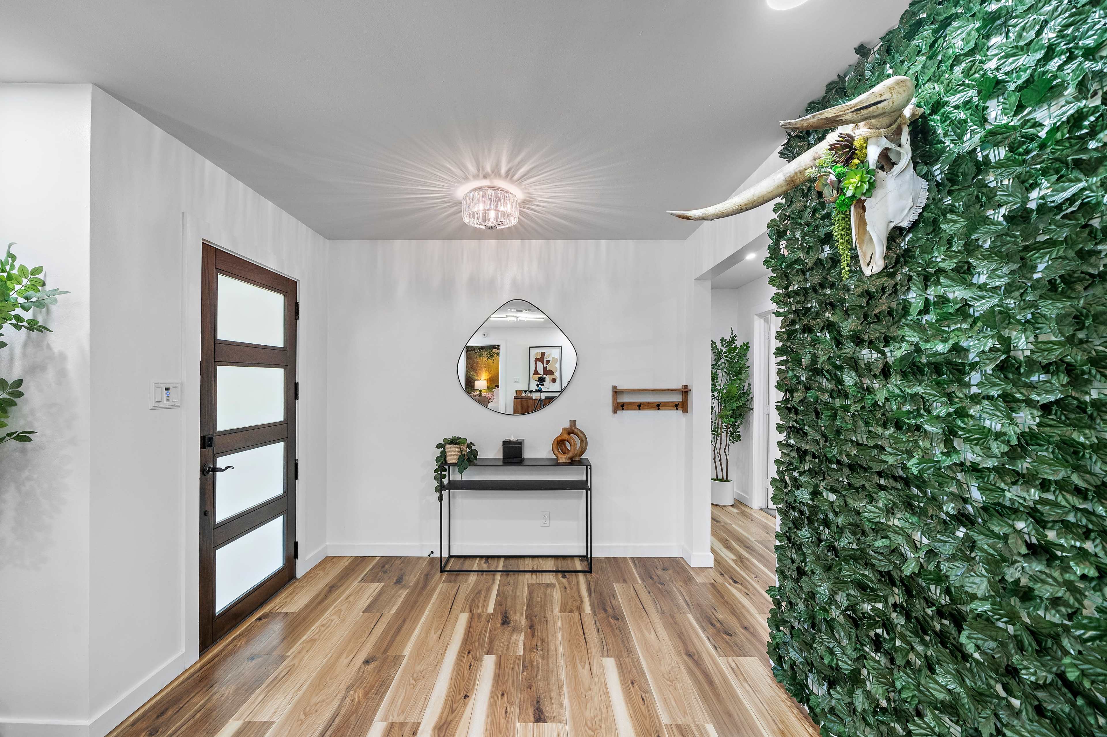 A modern entryway featuring a green leaf-covered wall, a decorative mirror above a table, and a door to the left.
