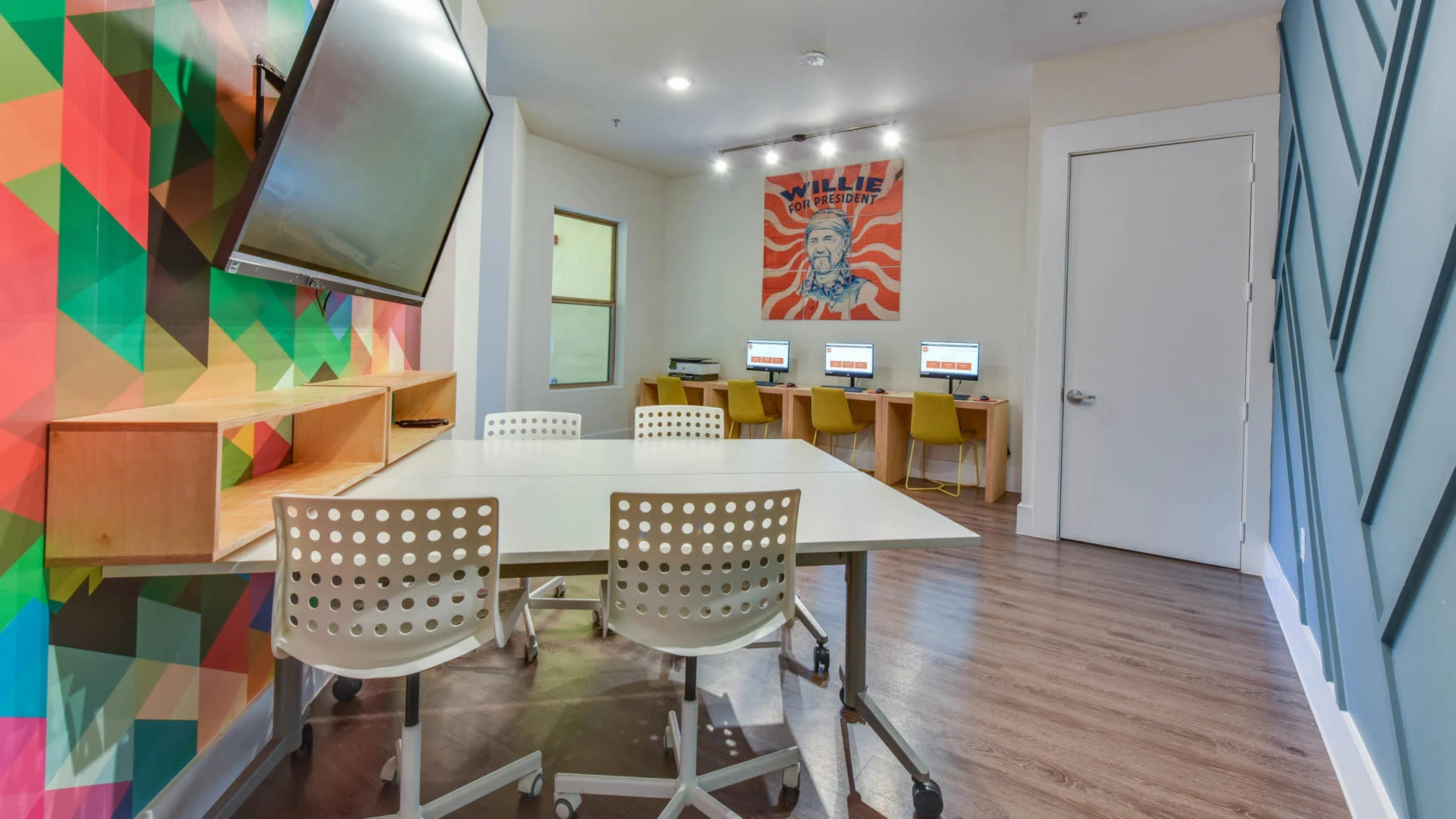The image shows a modern meeting room with a colorful geometric wall design, a large screen mounted on one side, and a table surrounded by four chairs, with computers set up against the back wall.