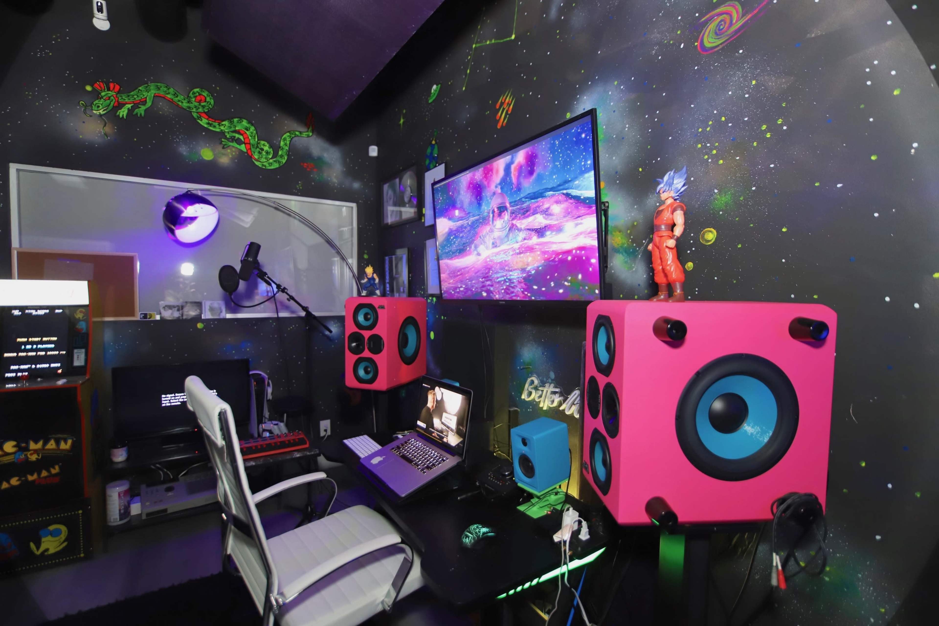 A colorful recording studio features pink speakers, a monitor displaying vibrant graphics, and a desk with a laptop and microphone.