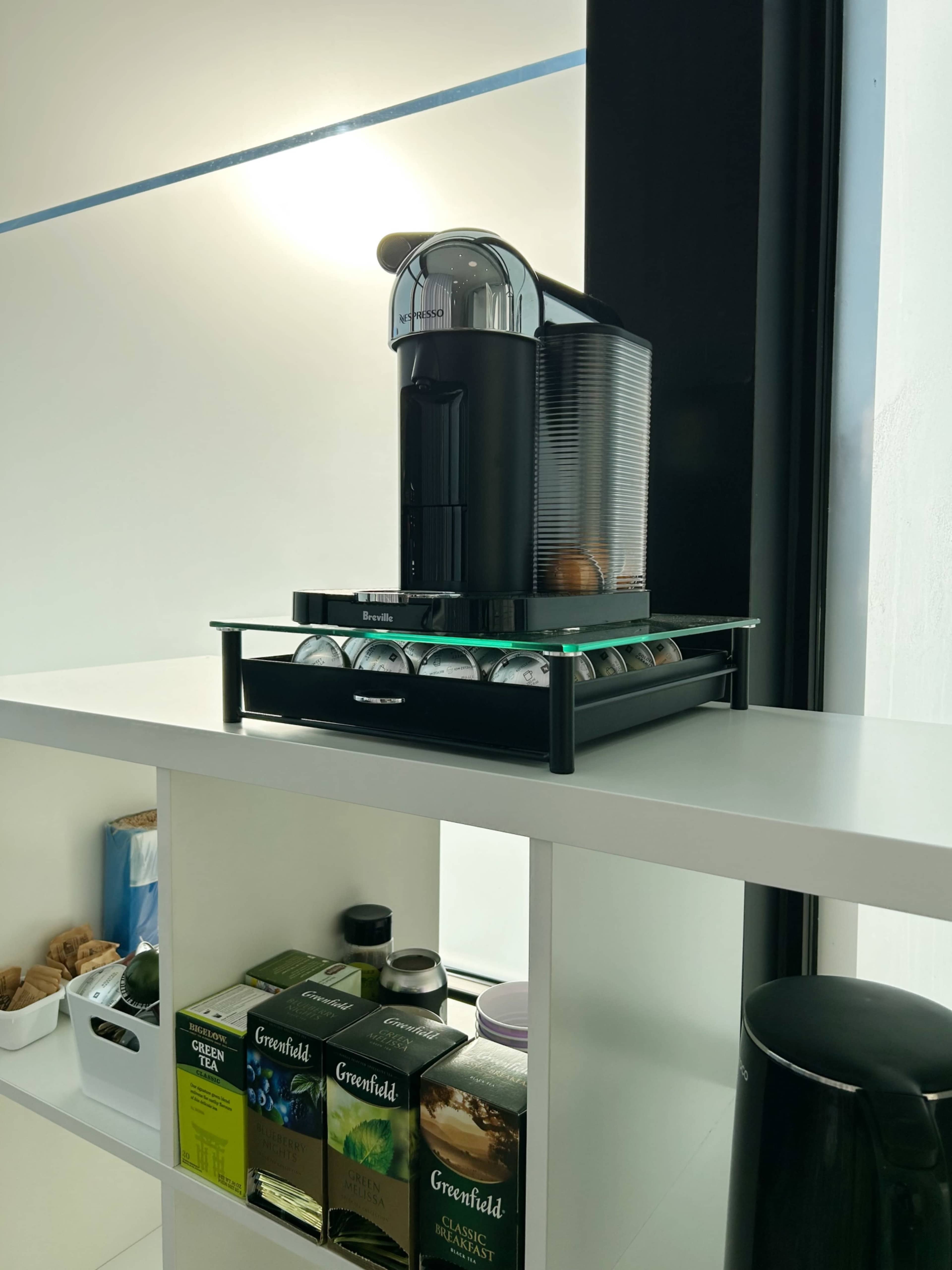 A coffee machine sits on a glass shelf above a neatly organized display of tea boxes and a container.