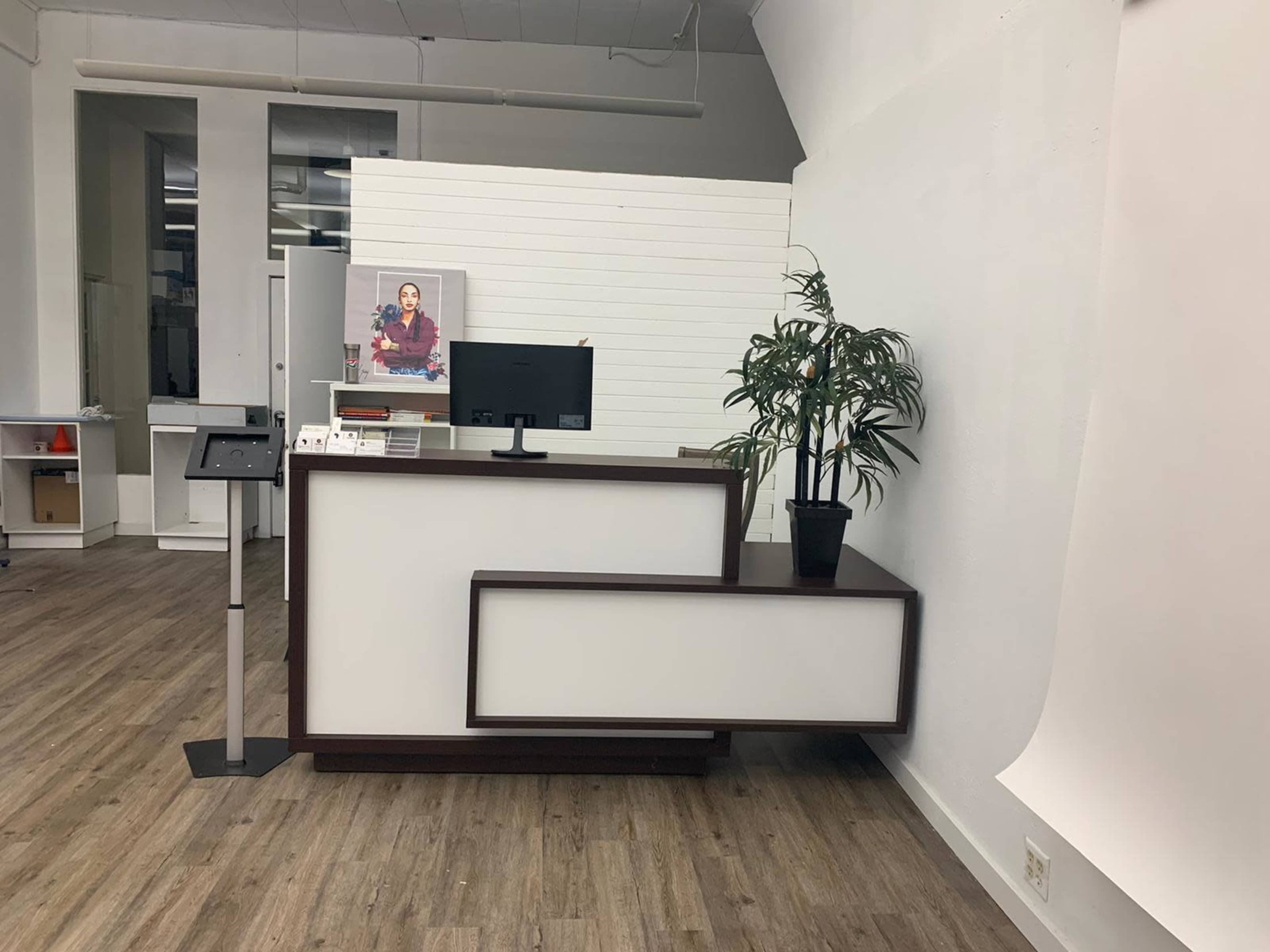 A clean reception desk with a monitor and a plant, set in a brightly lit, minimalist office space.