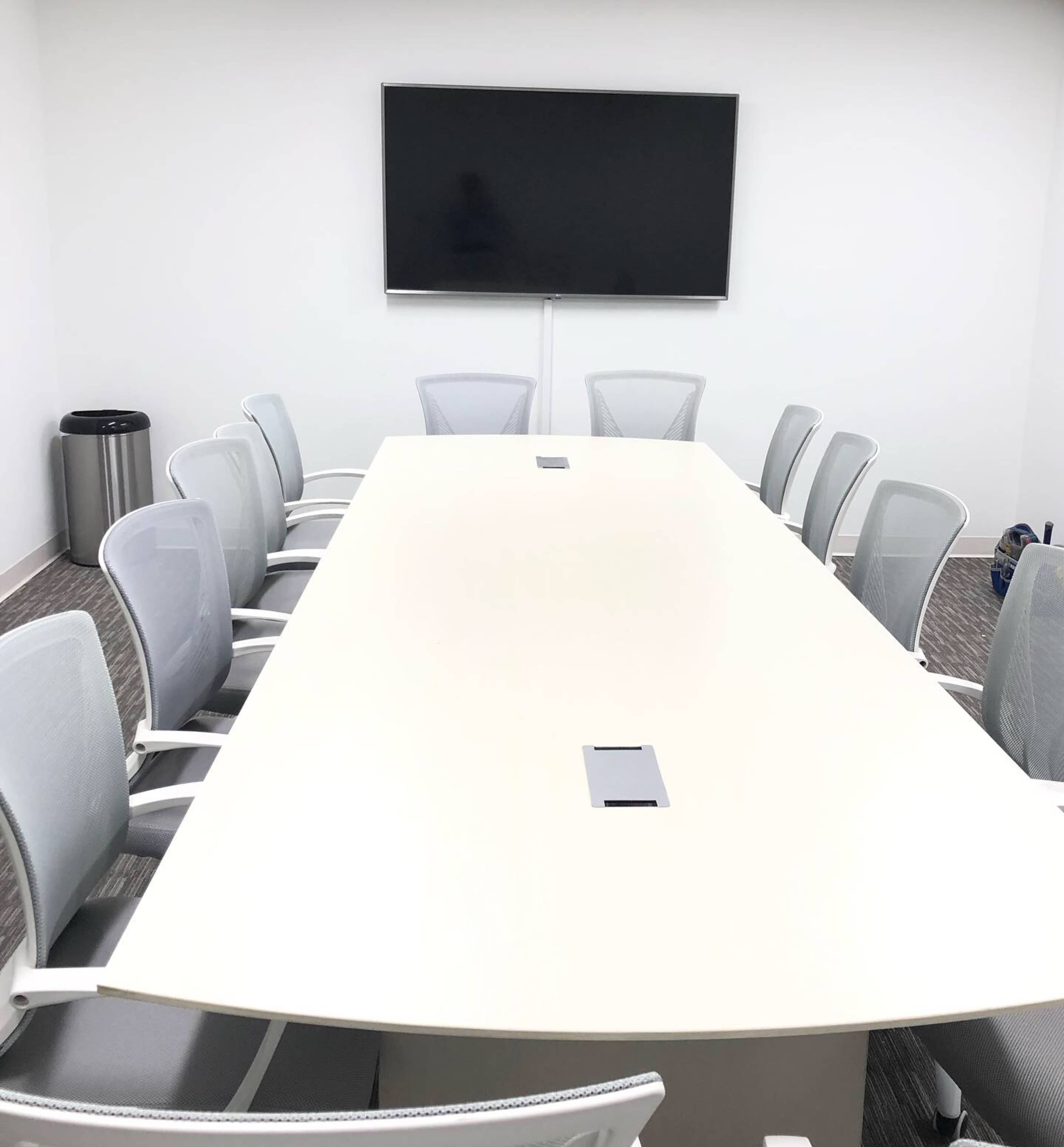 A brightly lit conference room features a long table surrounded by ergonomic chairs and a mounted television screen on the white wall.