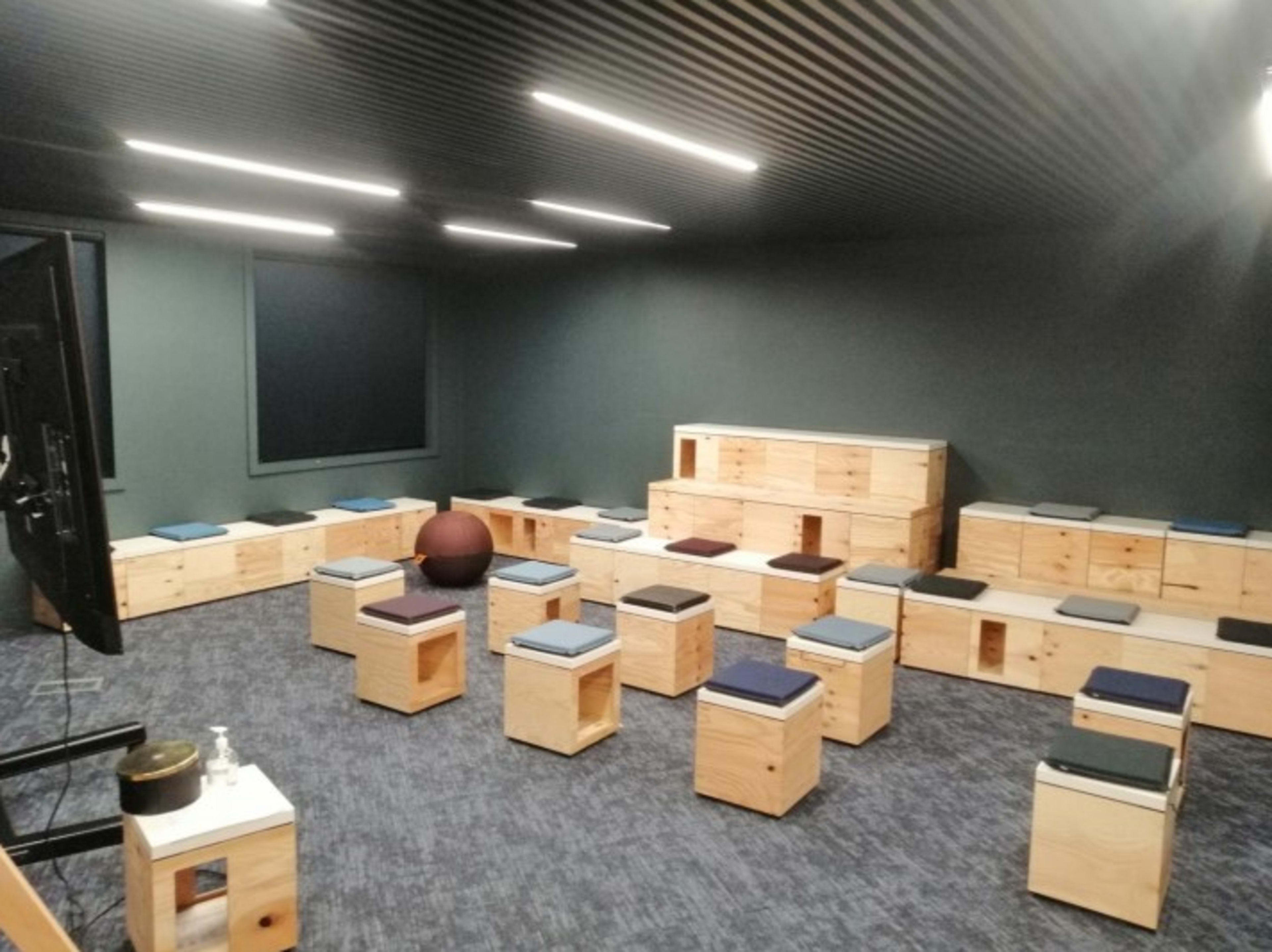 The image shows a modern, minimalistic room equipped with wooden seating and small tables arranged in a semi-circle, along with a large screen in one corner.