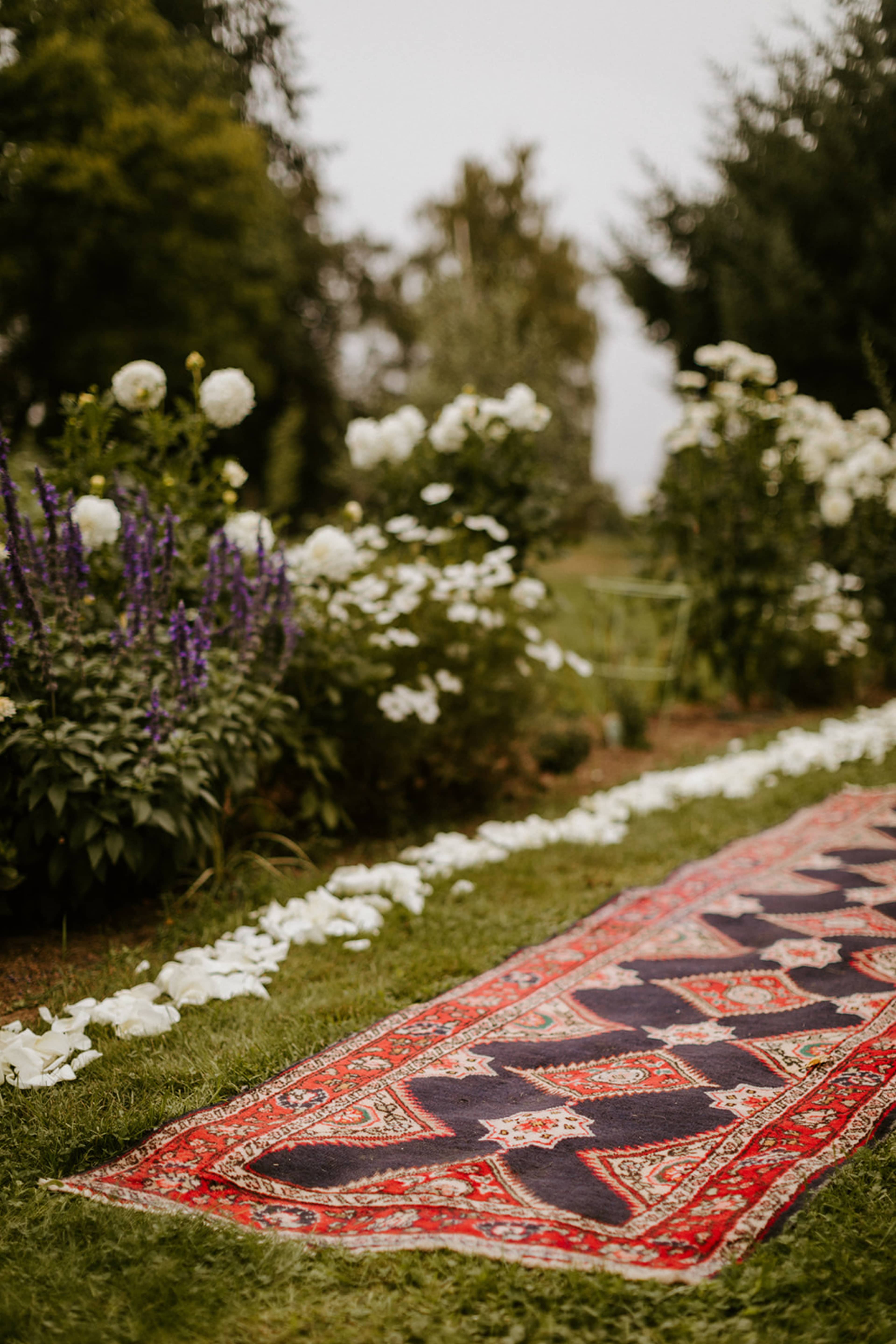 A colorful rug is placed on a grassy path lined with flower petals, surrounded by blooming plants and trees.