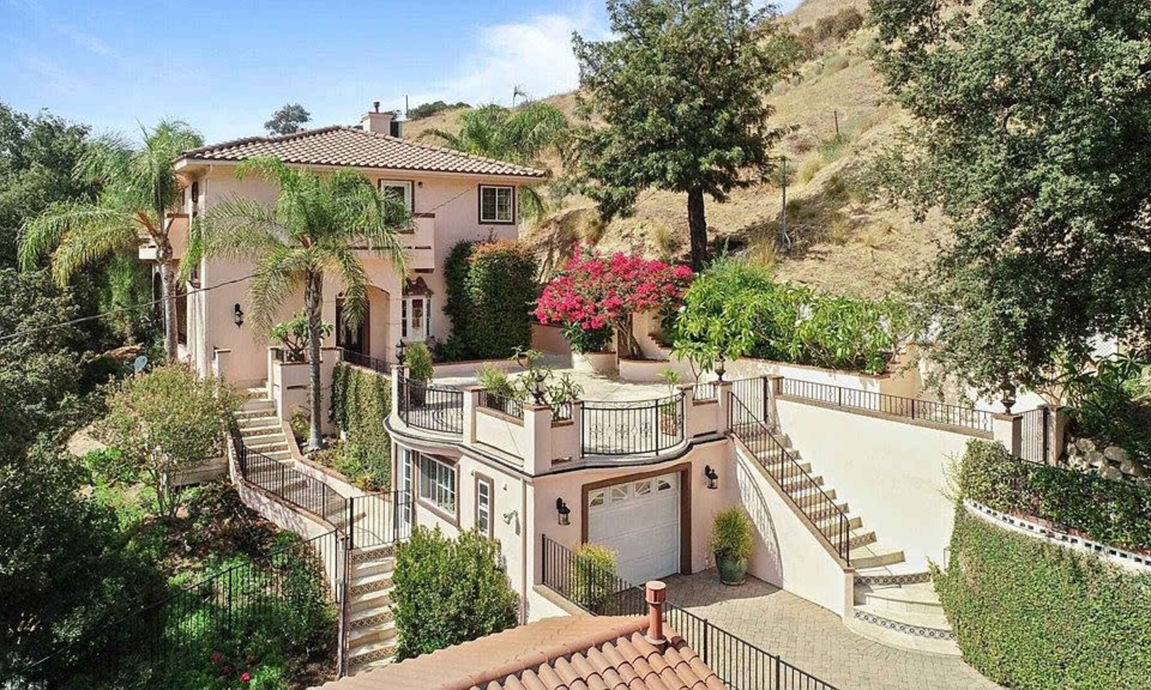 A two-story pink house with a tile roof, surrounded by landscaped gardens and palm trees, situated on a hillside.