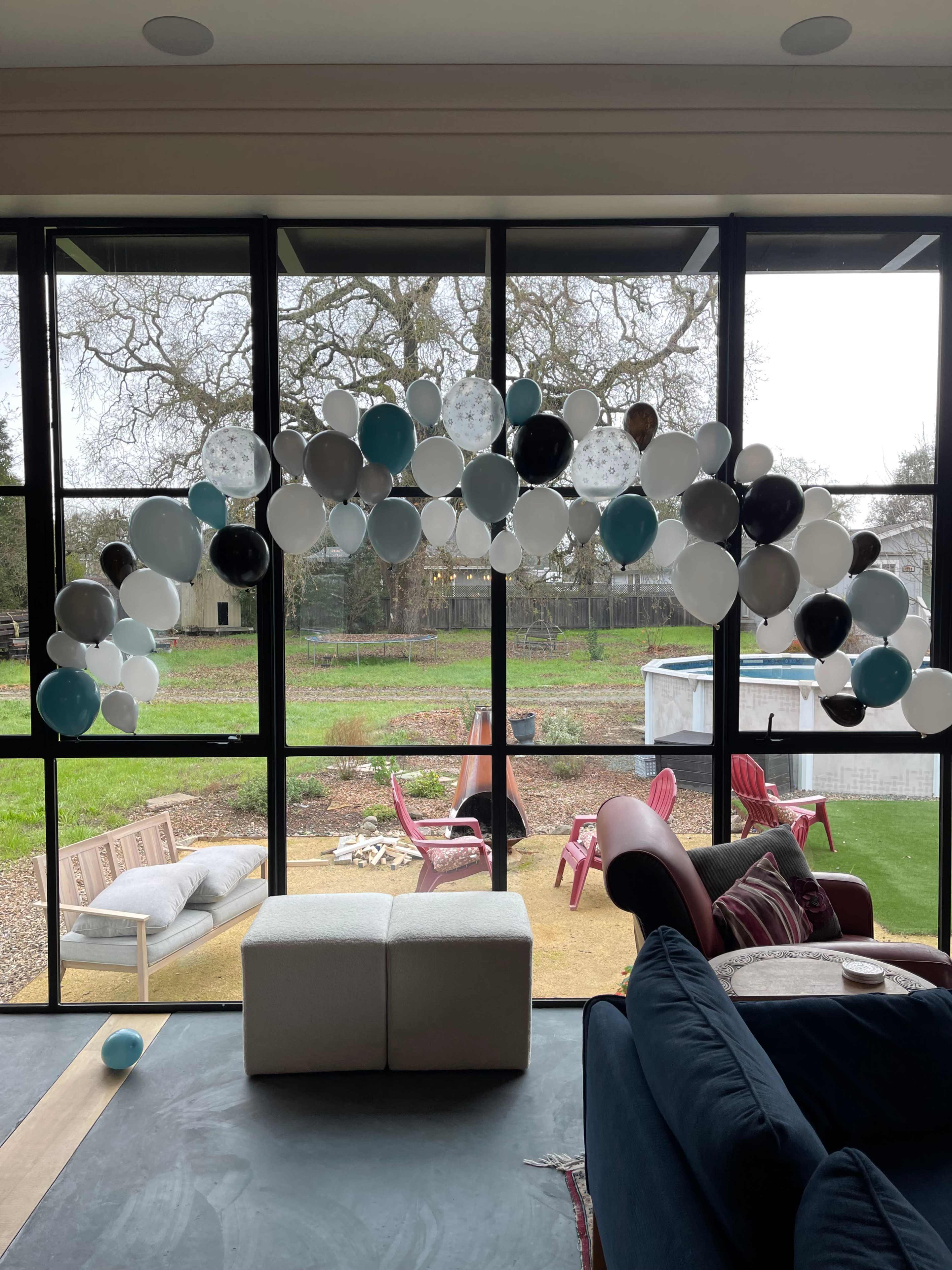 A decorative arch made of balloons in various colors is positioned in front of large glass windows overlooking a grassy yard with chairs.