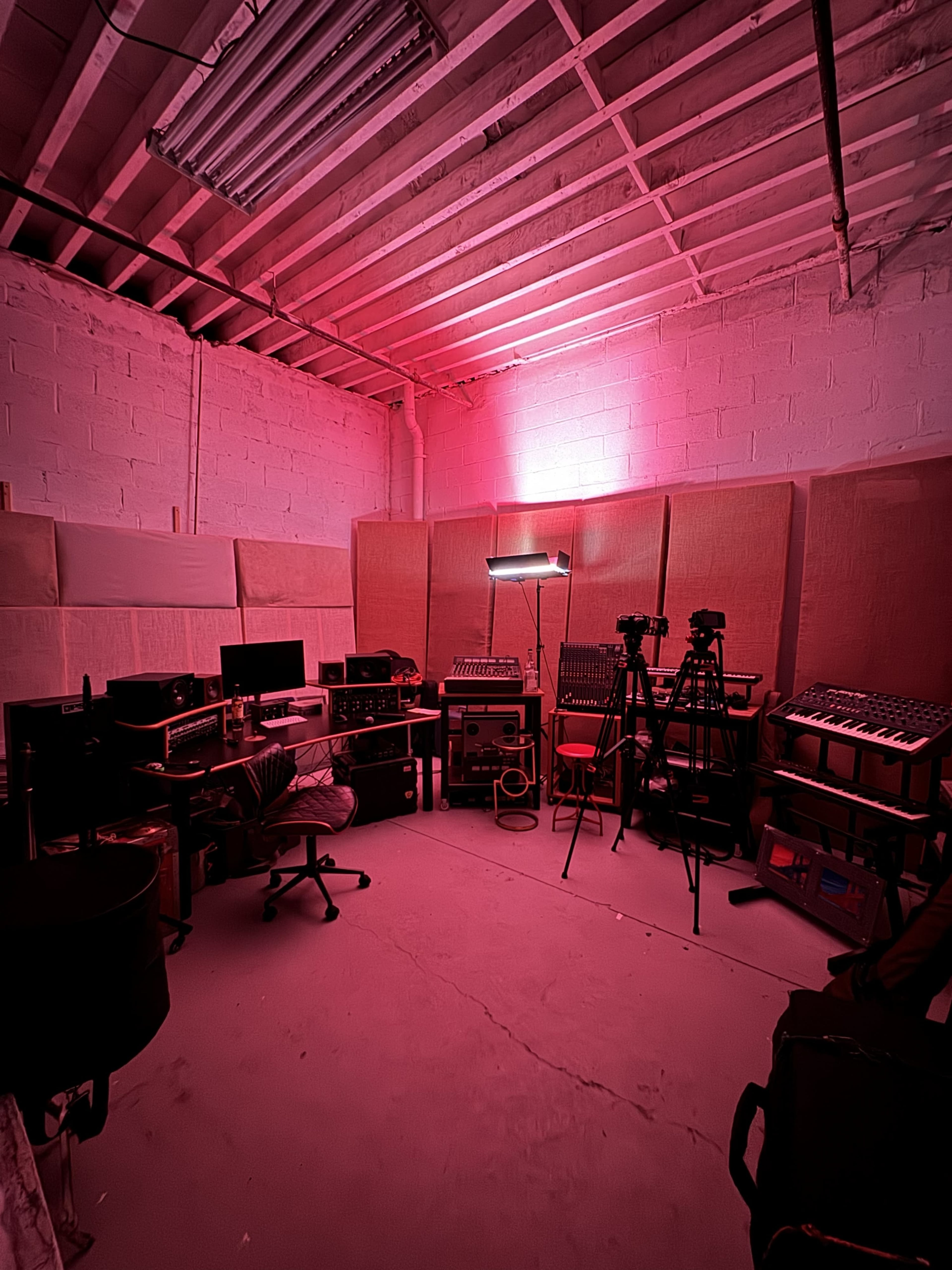The image shows a dimly lit music recording studio with various equipment, including computers, cameras, and synthesizers, arranged against the walls.