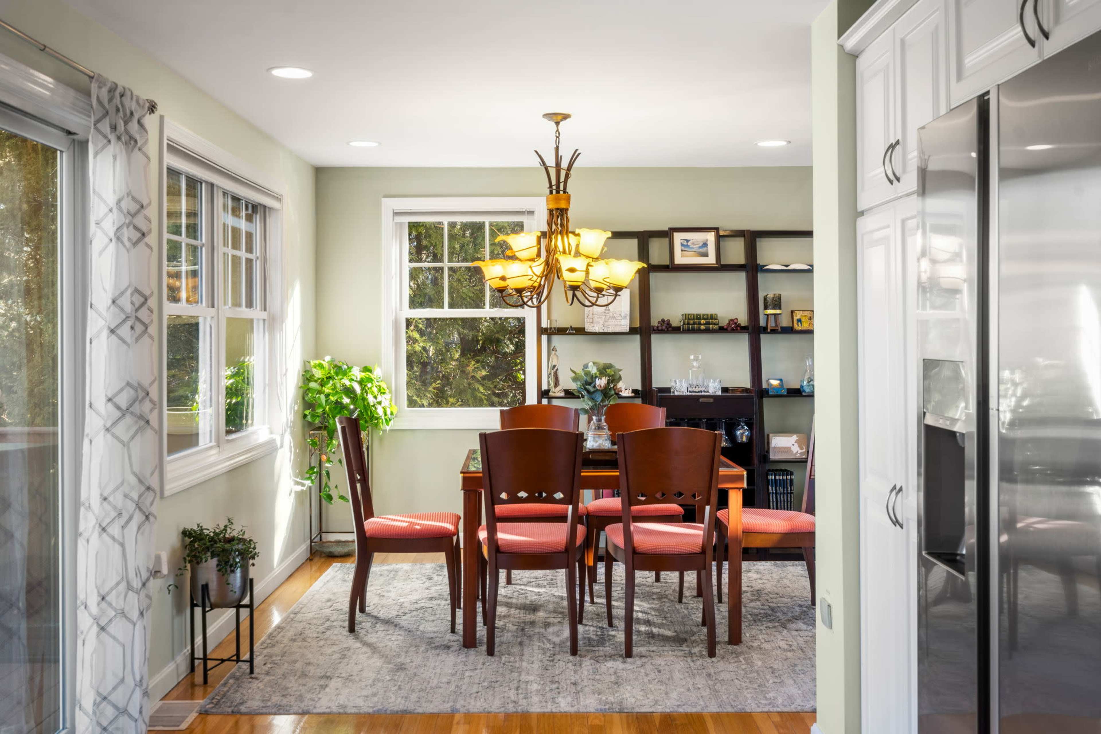 A dining area features a wooden table surrounded by four chairs, illuminated by a chandelier, with large windows and a shelf displaying decorative items.