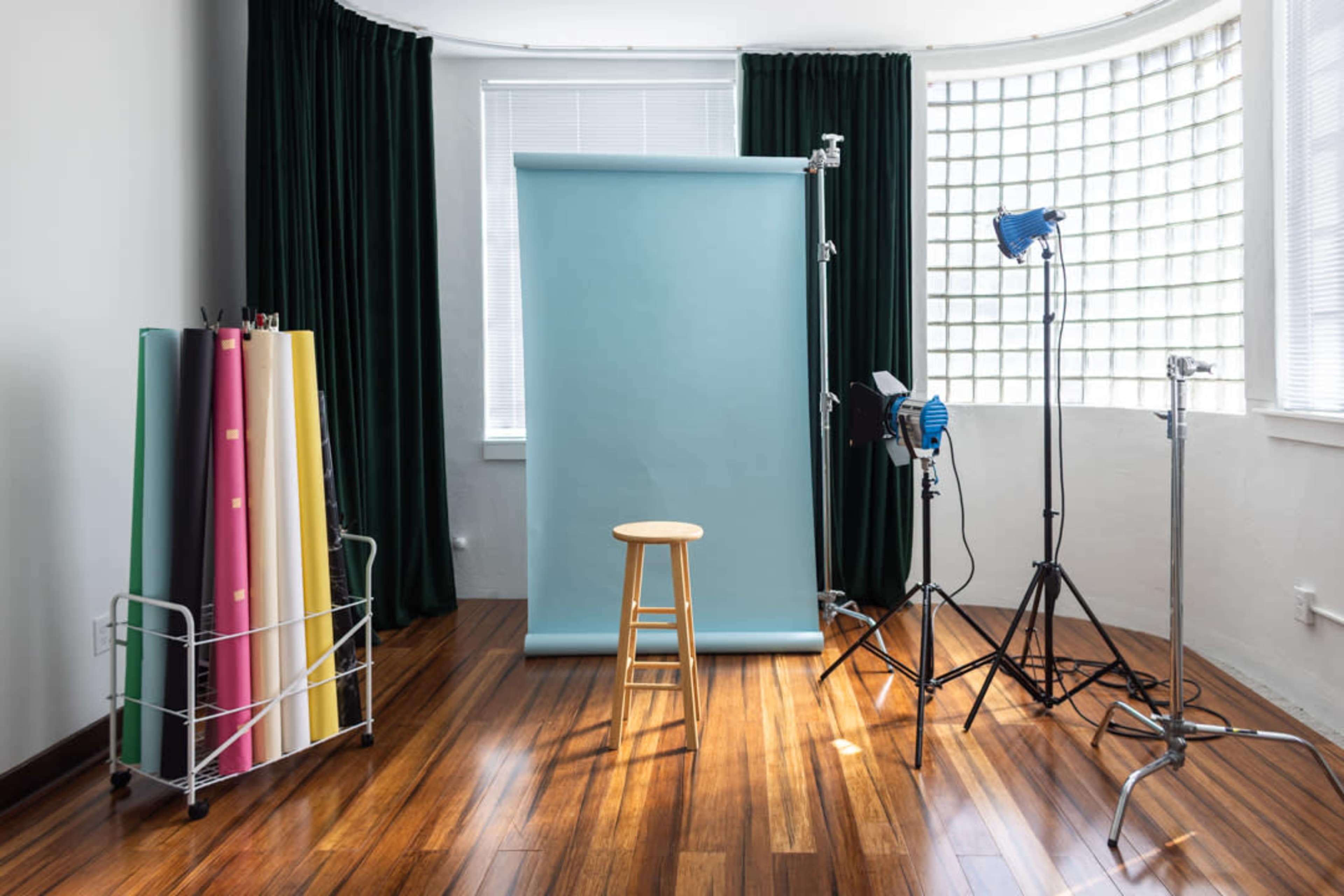 The image shows a photography studio with a wooden stool in front of a light blue backdrop, surrounded by colorful rolls of paper and studio lighting equipment.