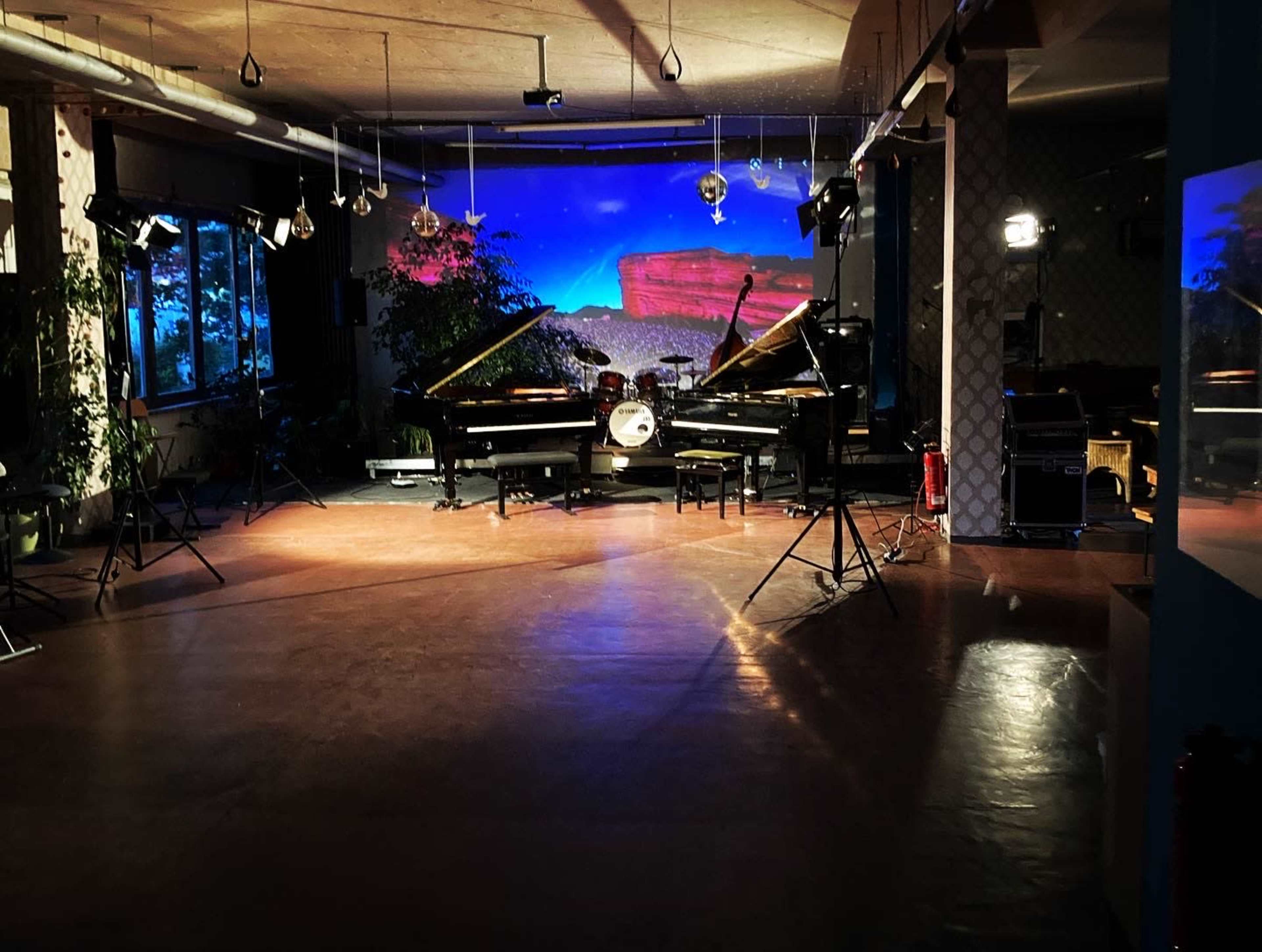 The image shows a spacious music venue featuring two grand pianos facing a drum set, with colorful lights and decorative plants in the background.