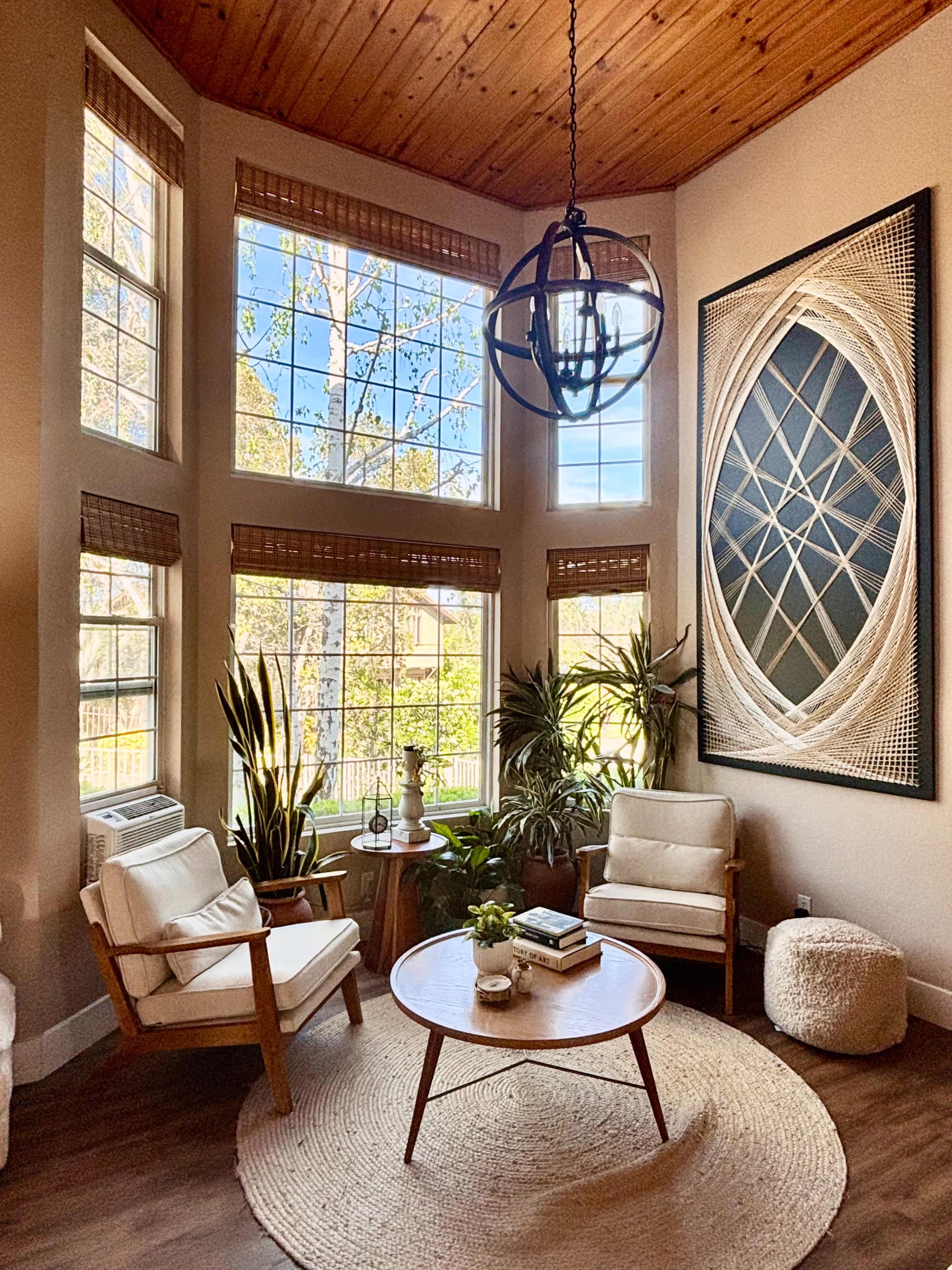 A cozy living room features large windows, a circular wooden coffee table, two armchairs, and various indoor plants under a wooden ceiling with a decorative light fixture.