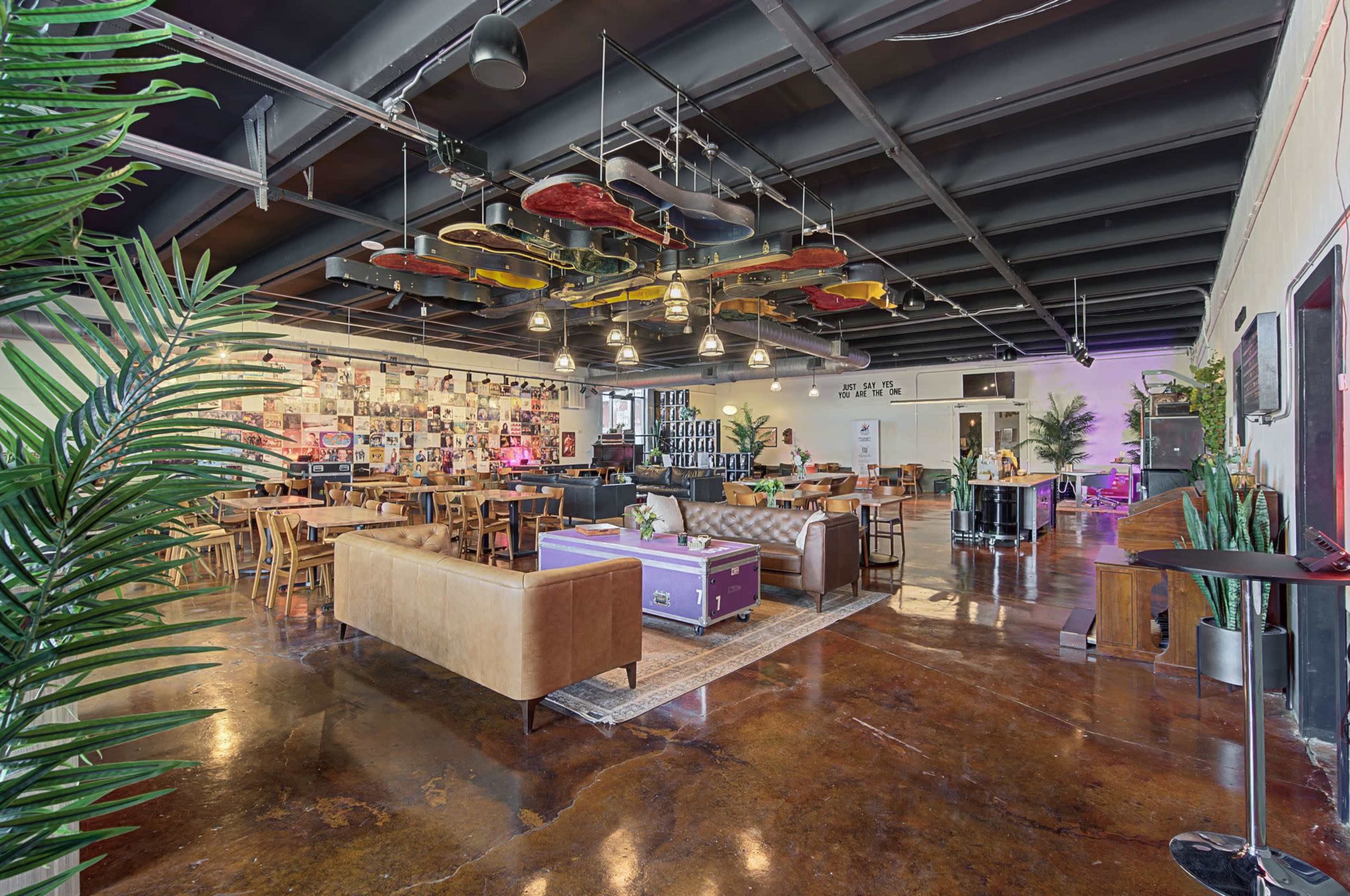 The image shows a spacious, modern lounge area featuring a mix of wooden furniture, colorful decor, large plants, and unique lighting fixtures.