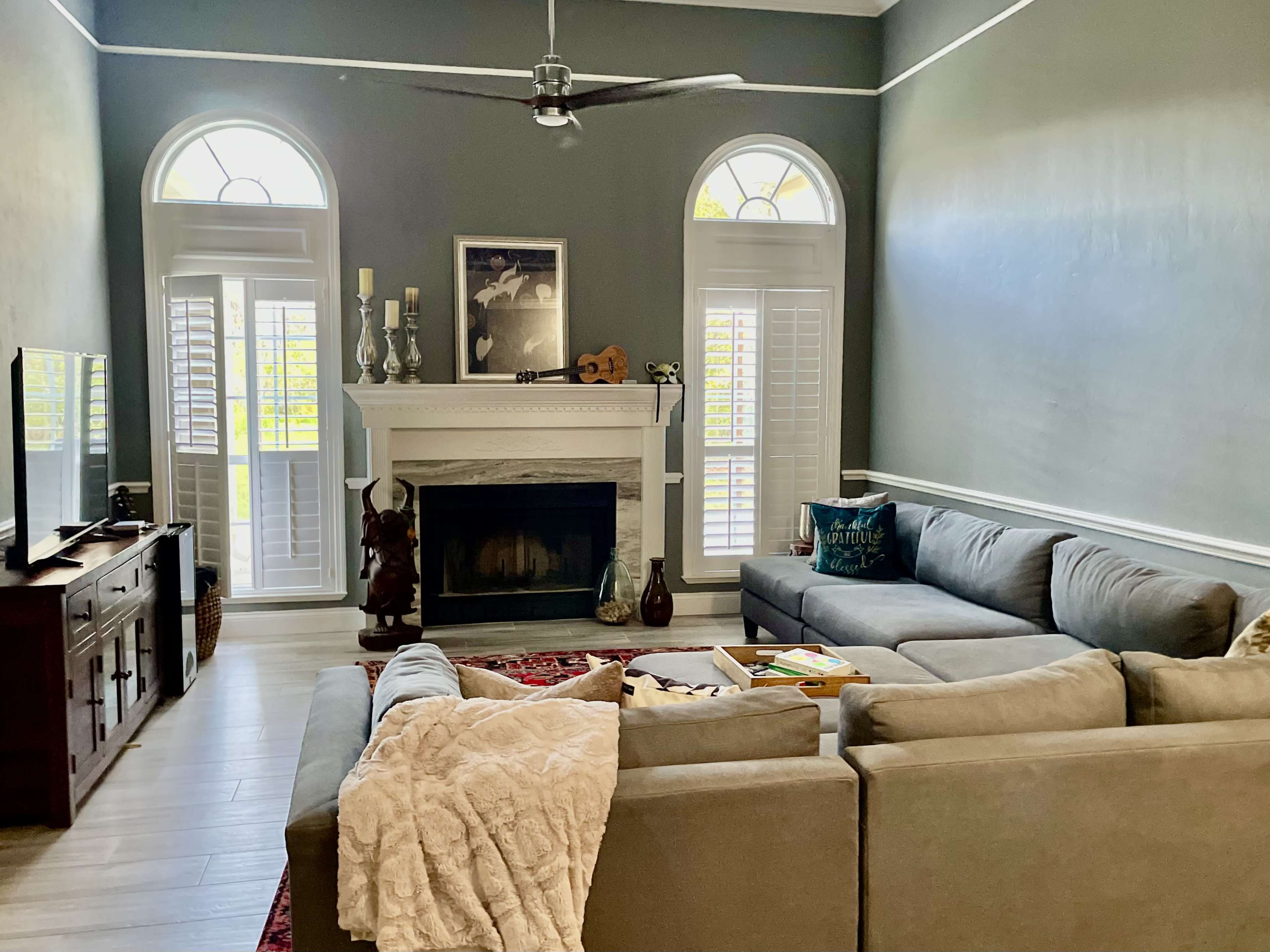 The living room features a large sectional sofa, a fireplace with decorative items, and tall windows with shutters.