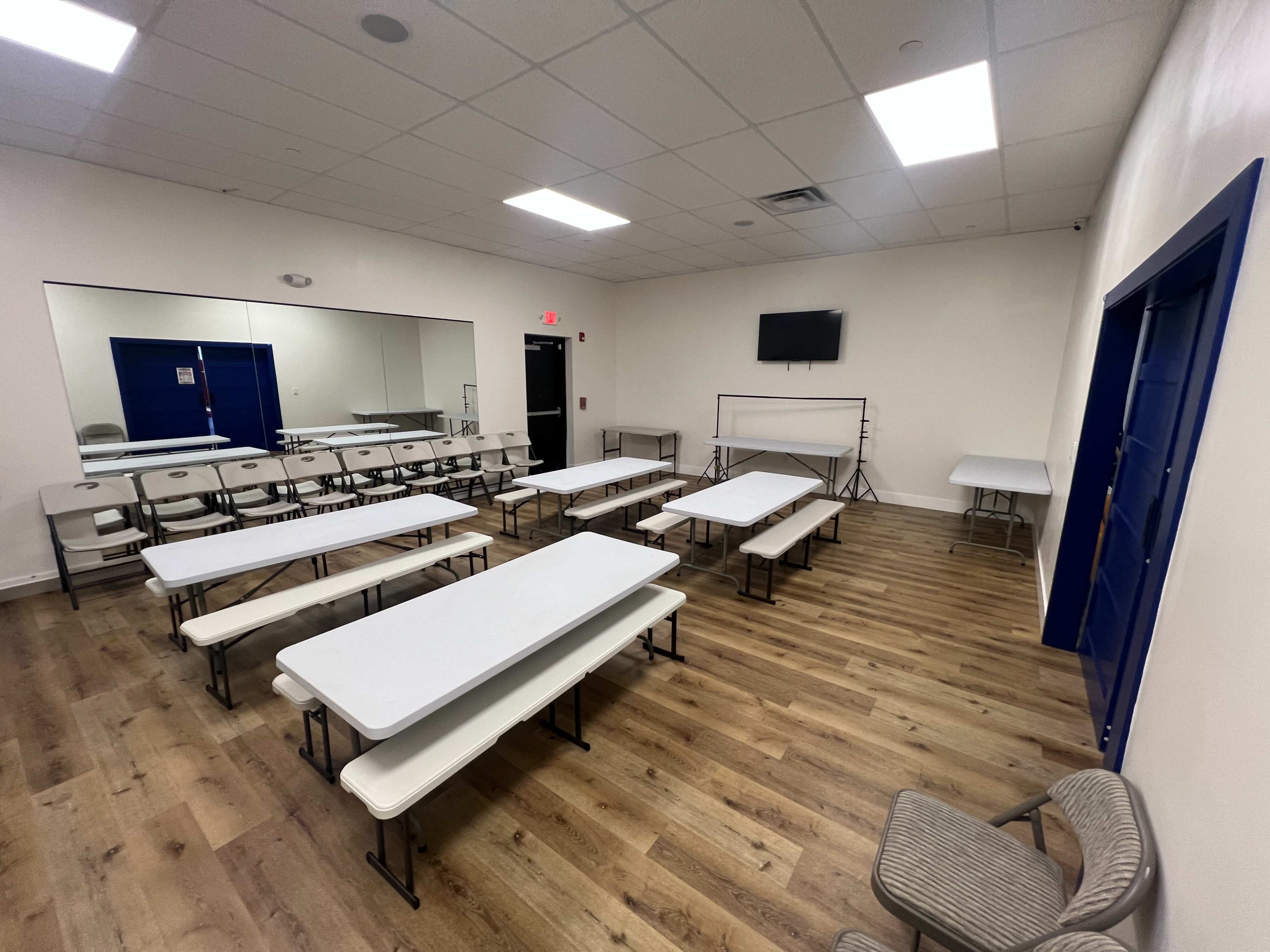 The image shows a spacious meeting room furnished with multiple rectangular tables and chairs arranged in rows, with a large mirror and a television mounted on the wall.