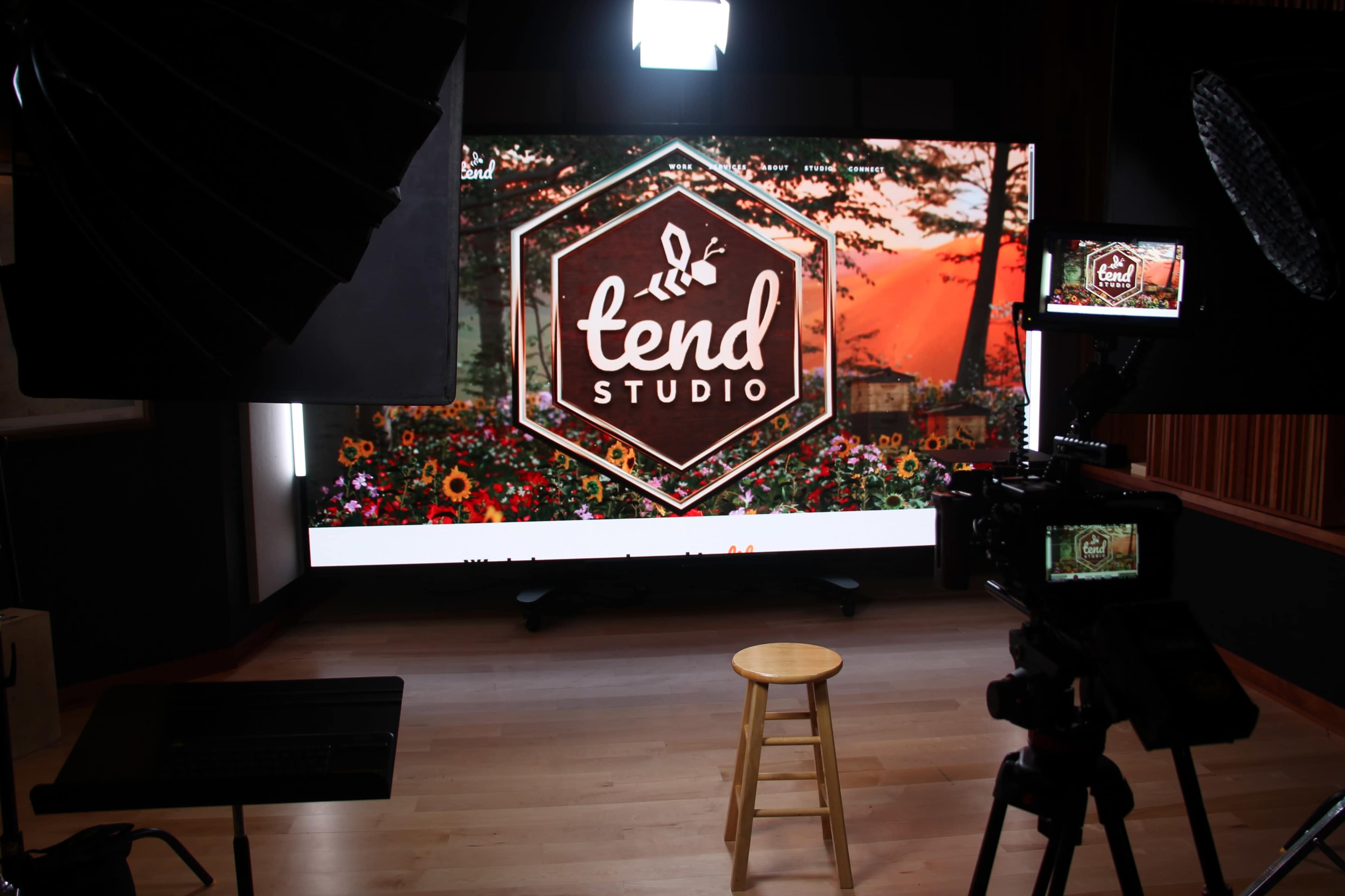 The image shows a studio setup featuring a large screen displaying a logo with the words "tend STUDIO" and a backdrop of flowers and a sunset, with a stool positioned in front.