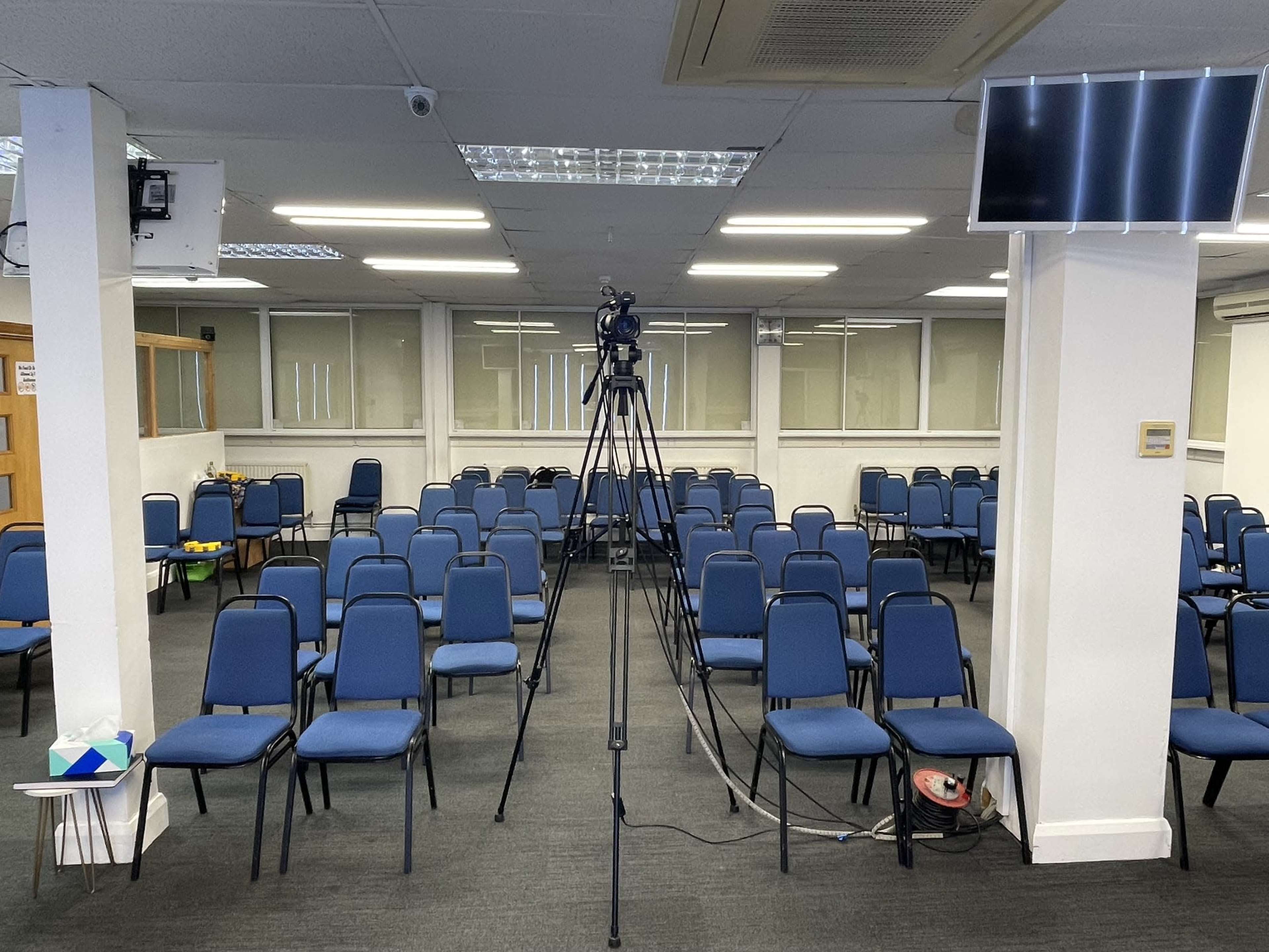 A large room filled with blue chairs arranged in rows, centered around a camera mounted on a tripod.