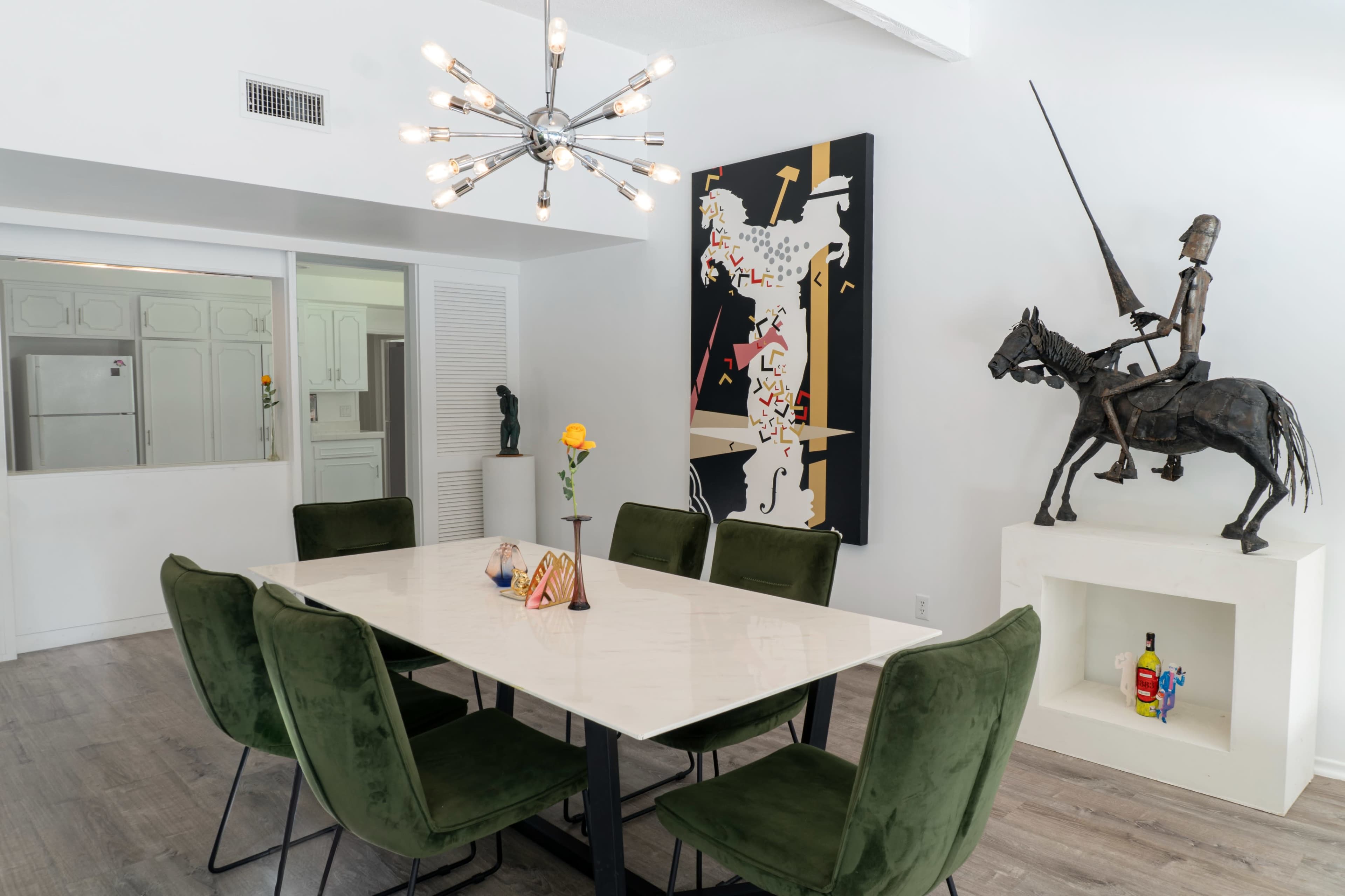 A modern dining area features a large marble table surrounded by green upholstered chairs, with a striking artwork and a statue of a knight on horseback displayed nearby.