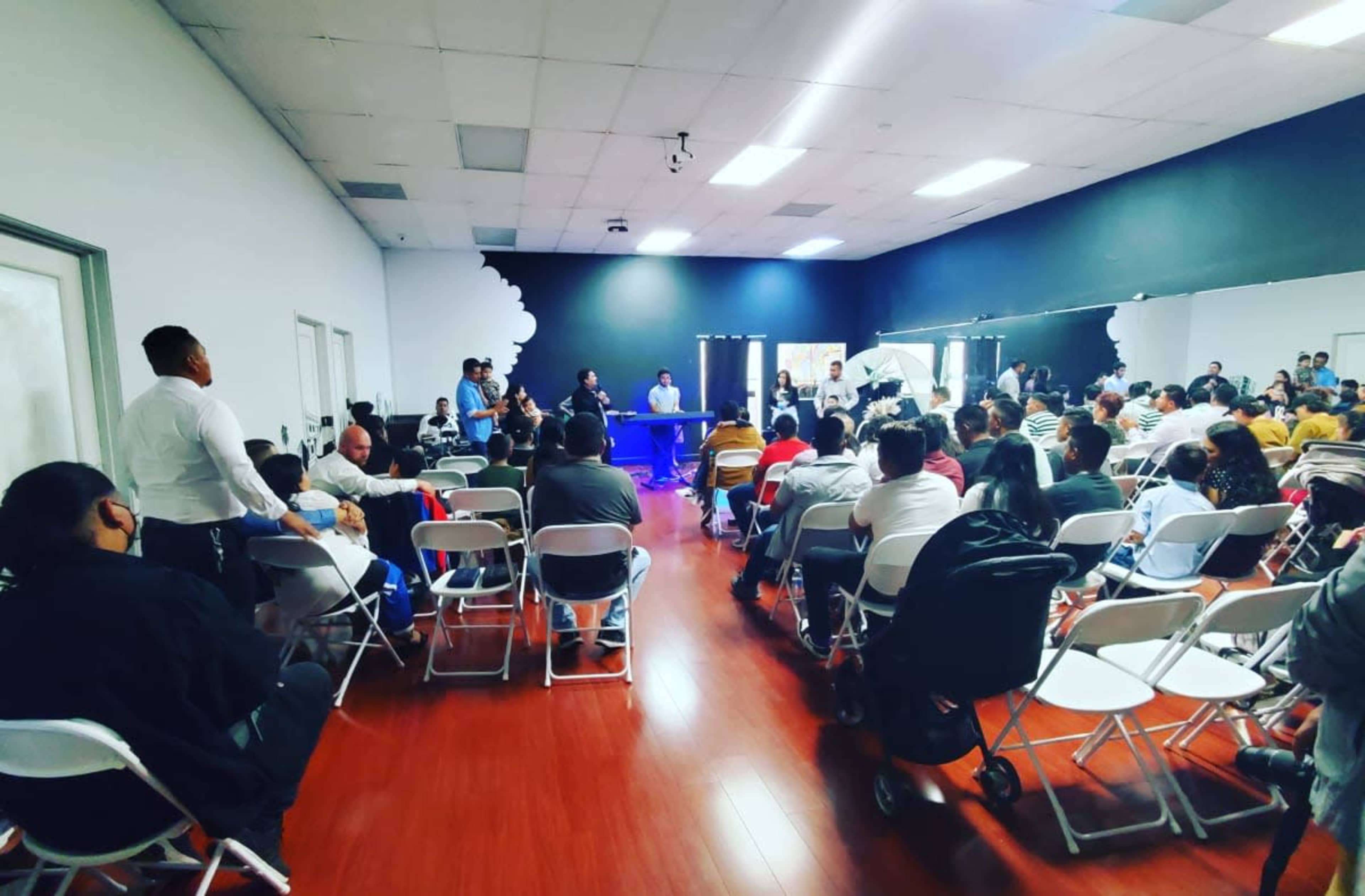 A large group of people is seated in white chairs, facing a panel at the front of a spacious, well-lit room.