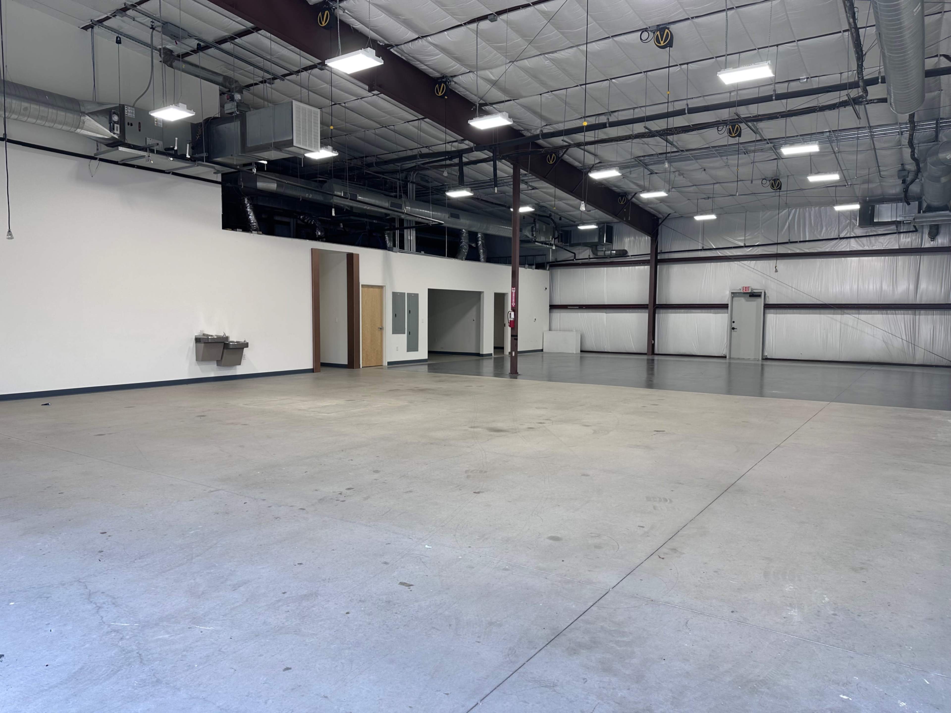 The image shows a spacious, empty industrial building interior with high ceilings, concrete flooring, and a few fixtures along the walls.