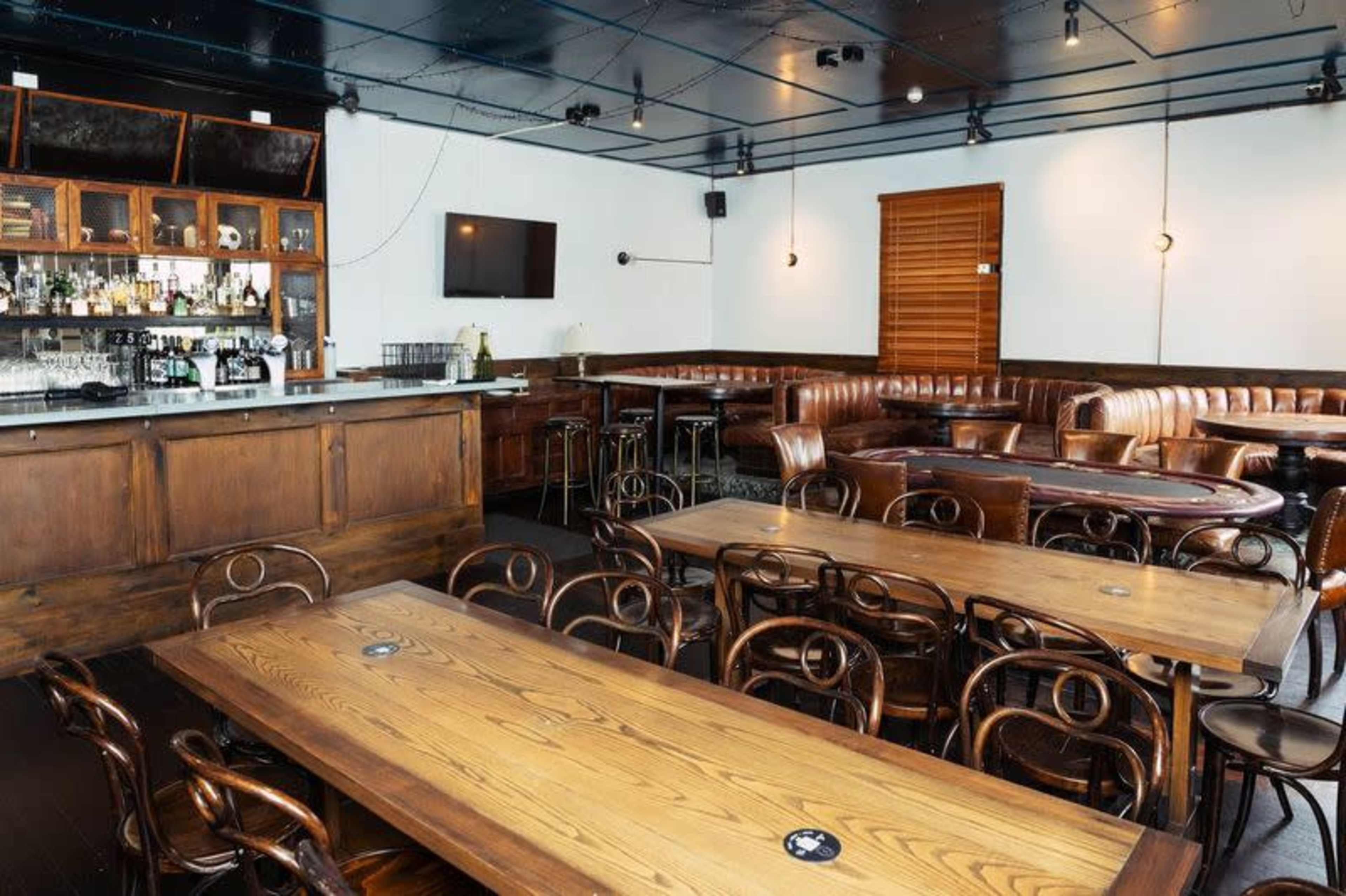 The image shows a modern bar interior with wooden tables, a bar area with shelves, and leather booth seating.