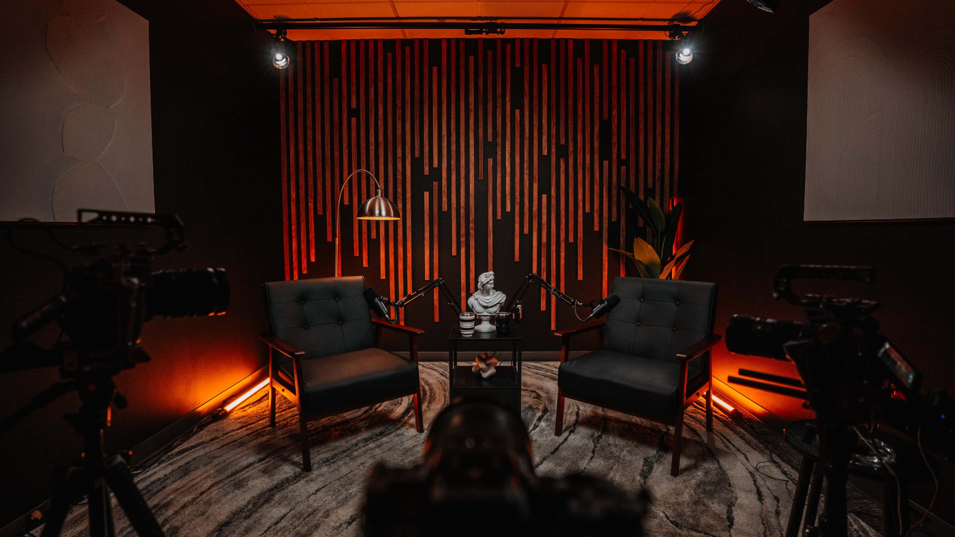 A recording studio features two chairs facing a central table, surrounded by cameras and illuminated by orange lighting against a striped wall backdrop.