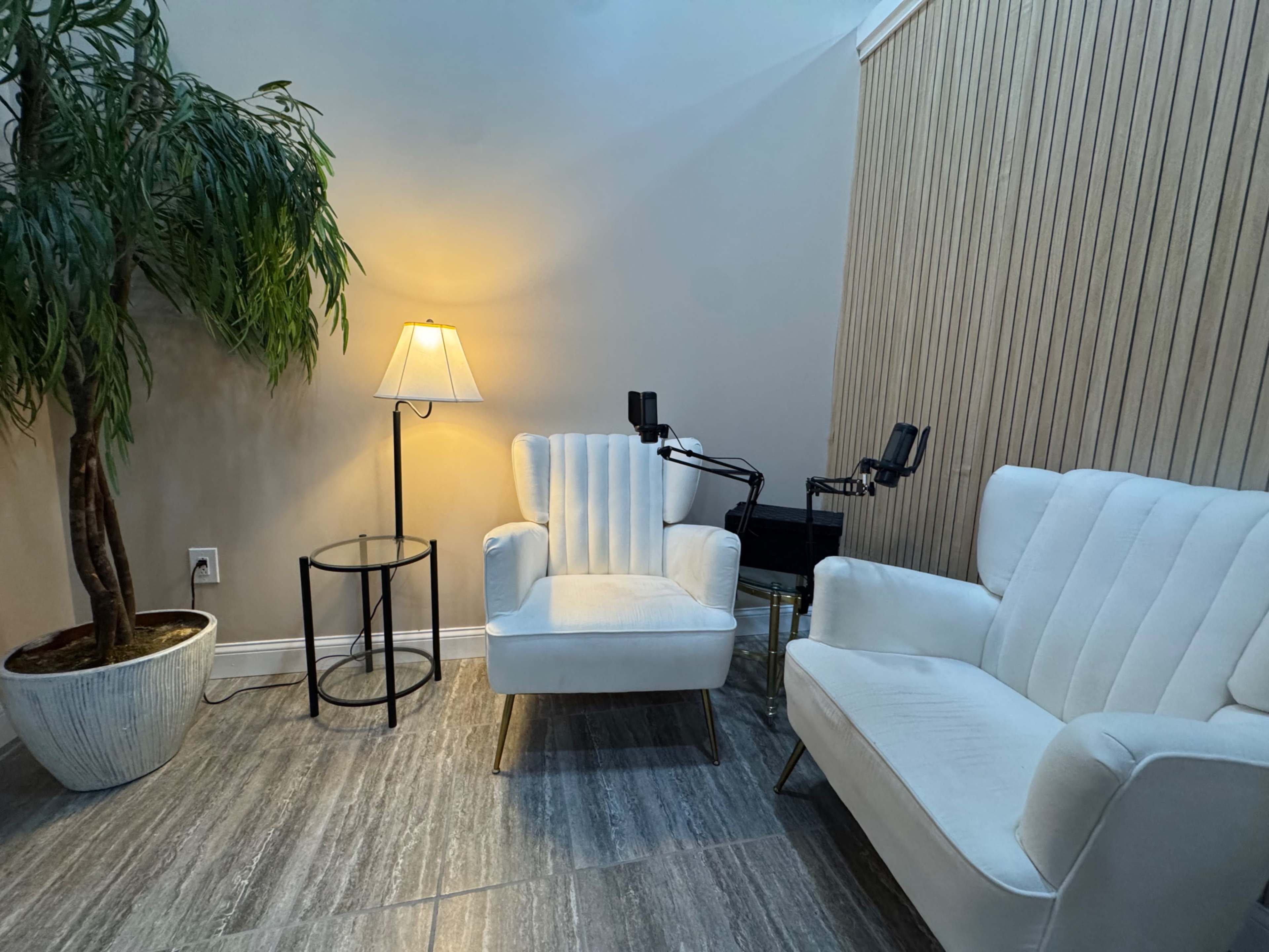 The image shows a corner of a room with two white chairs, a small round side table, a potted plant, and a lamp providing soft light.
