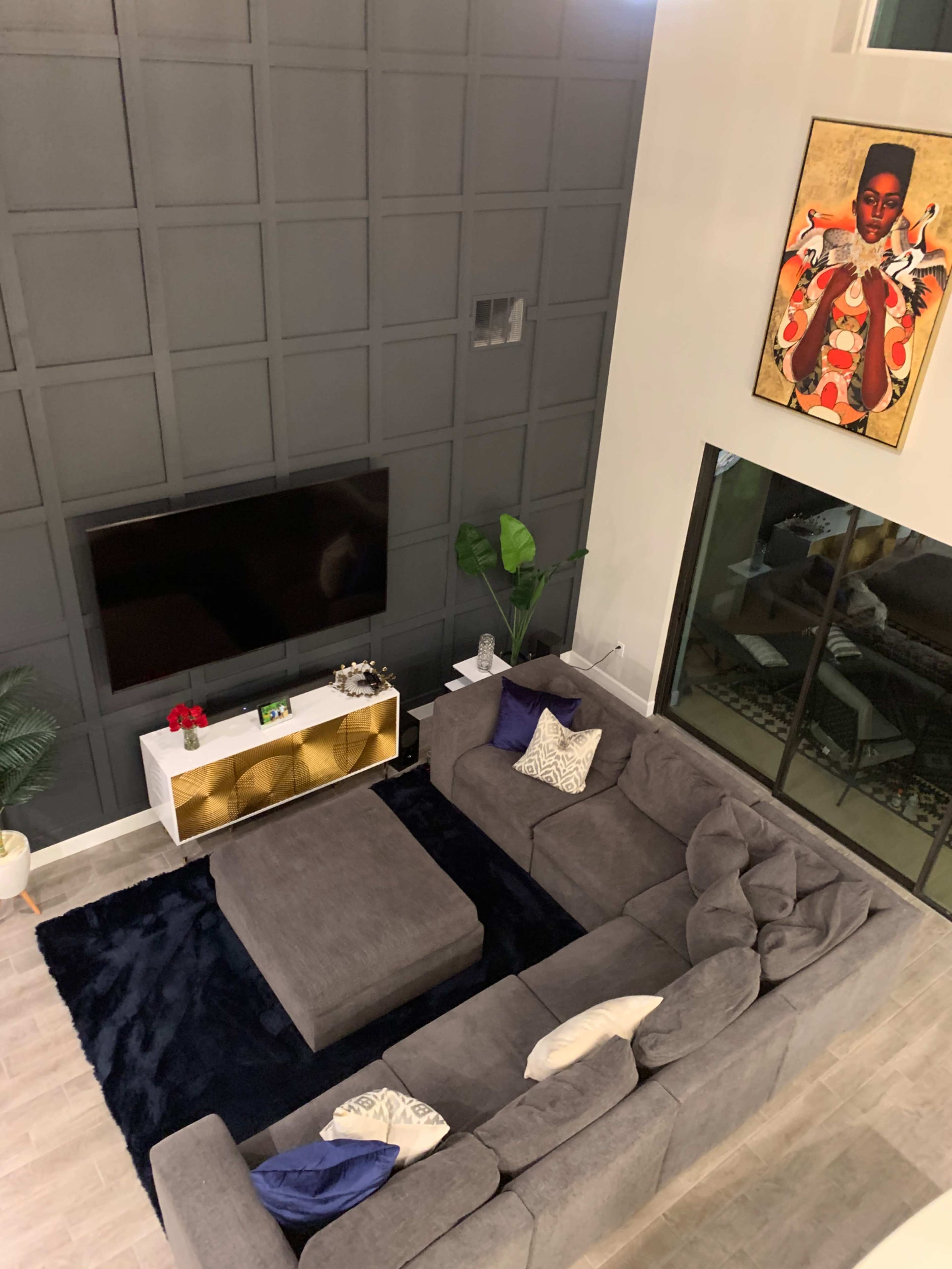 A modern living room with a large gray sectional sofa, a black area rug, a flat-screen TV mounted on a dark paneled wall, and a gold-accented console table with decorative items.