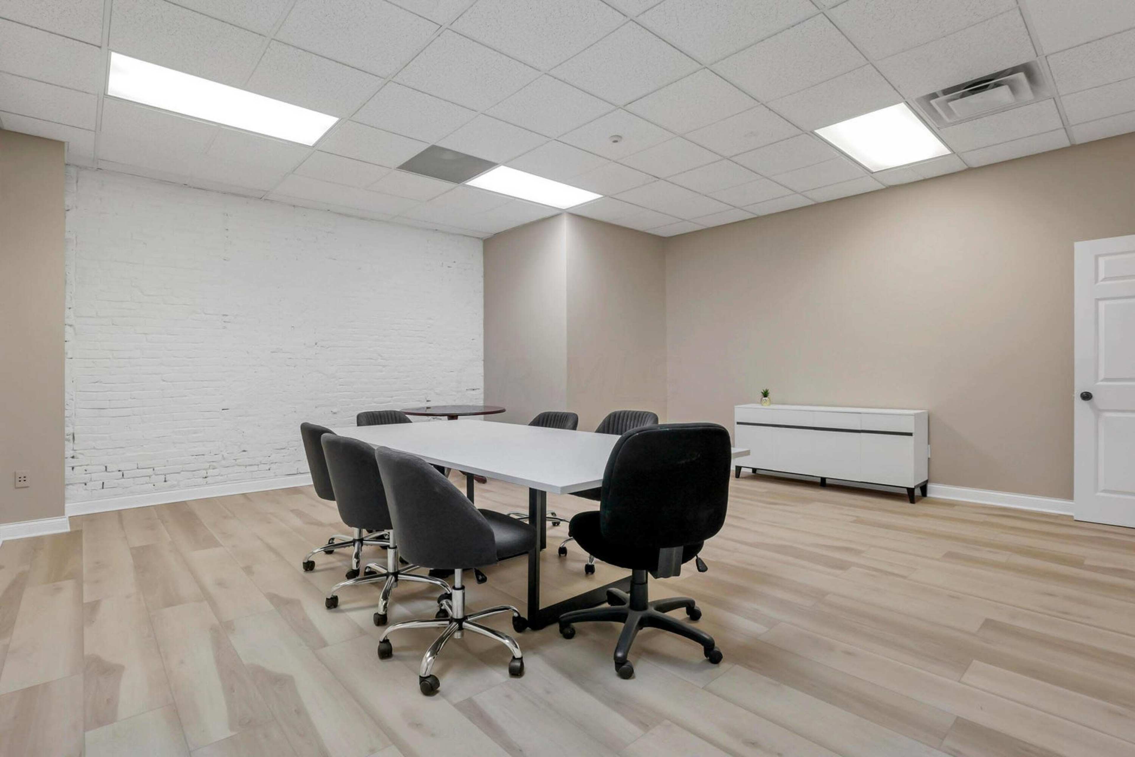 A conference room features a rectangular table surrounded by six chairs, with a white brick wall and a cabinet in the background.