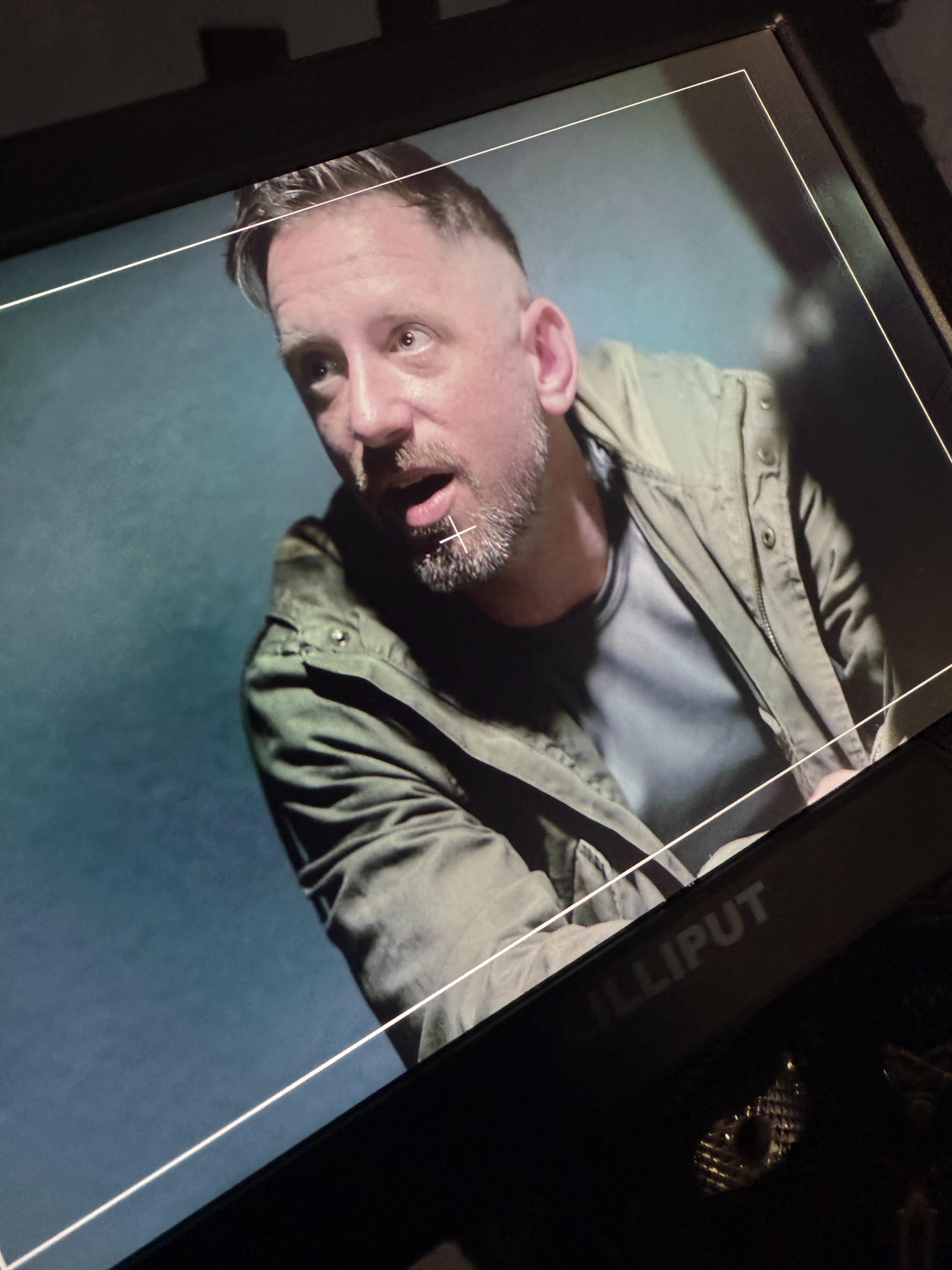 A man with short hair and a beard looks intently at something off camera while sitting in front of a monitor.