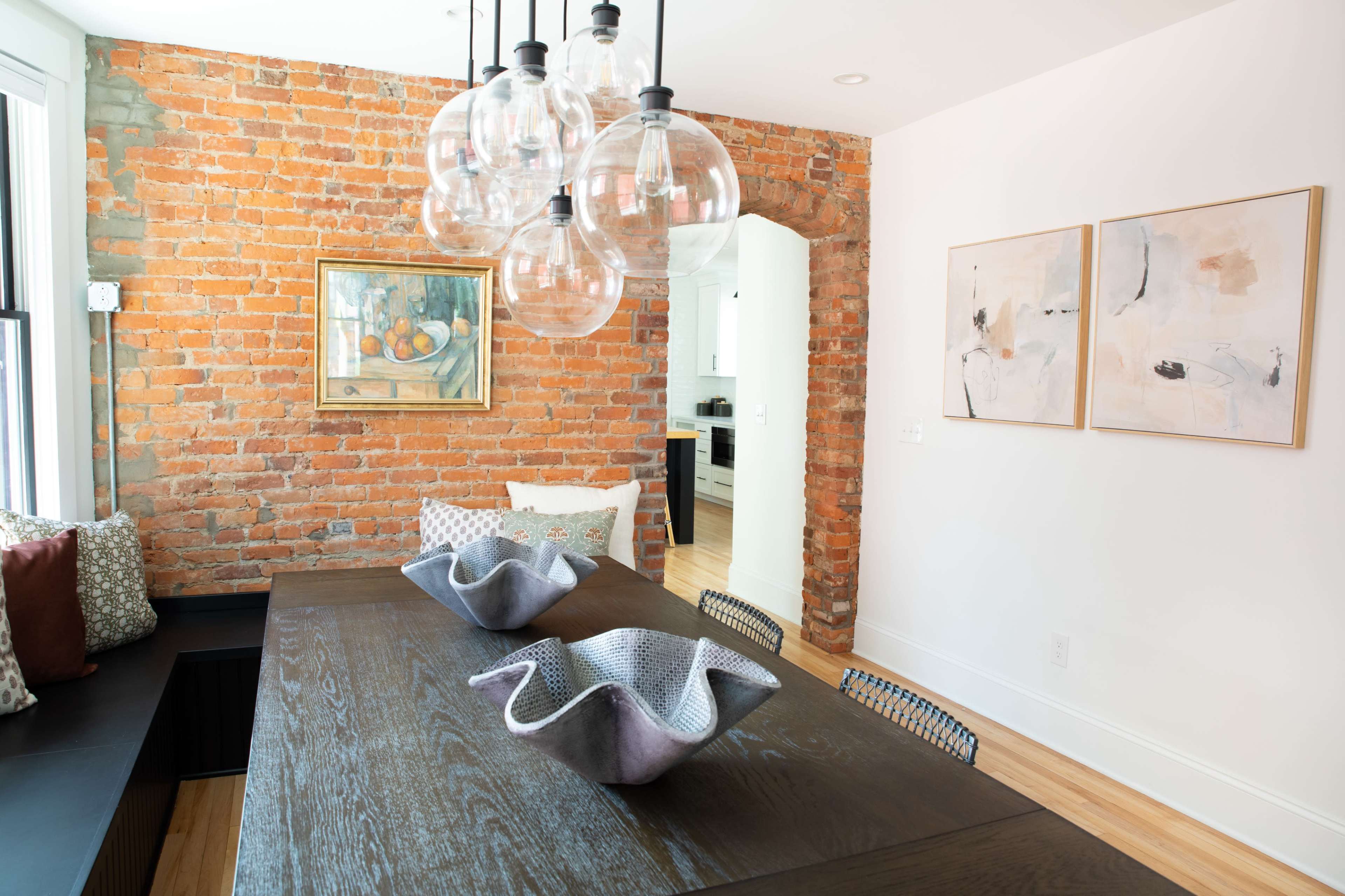 A dining area featuring a wooden table with decorative bowls, surrounded by a set of chairs, against a backdrop of exposed brick walls and modern light fixtures.