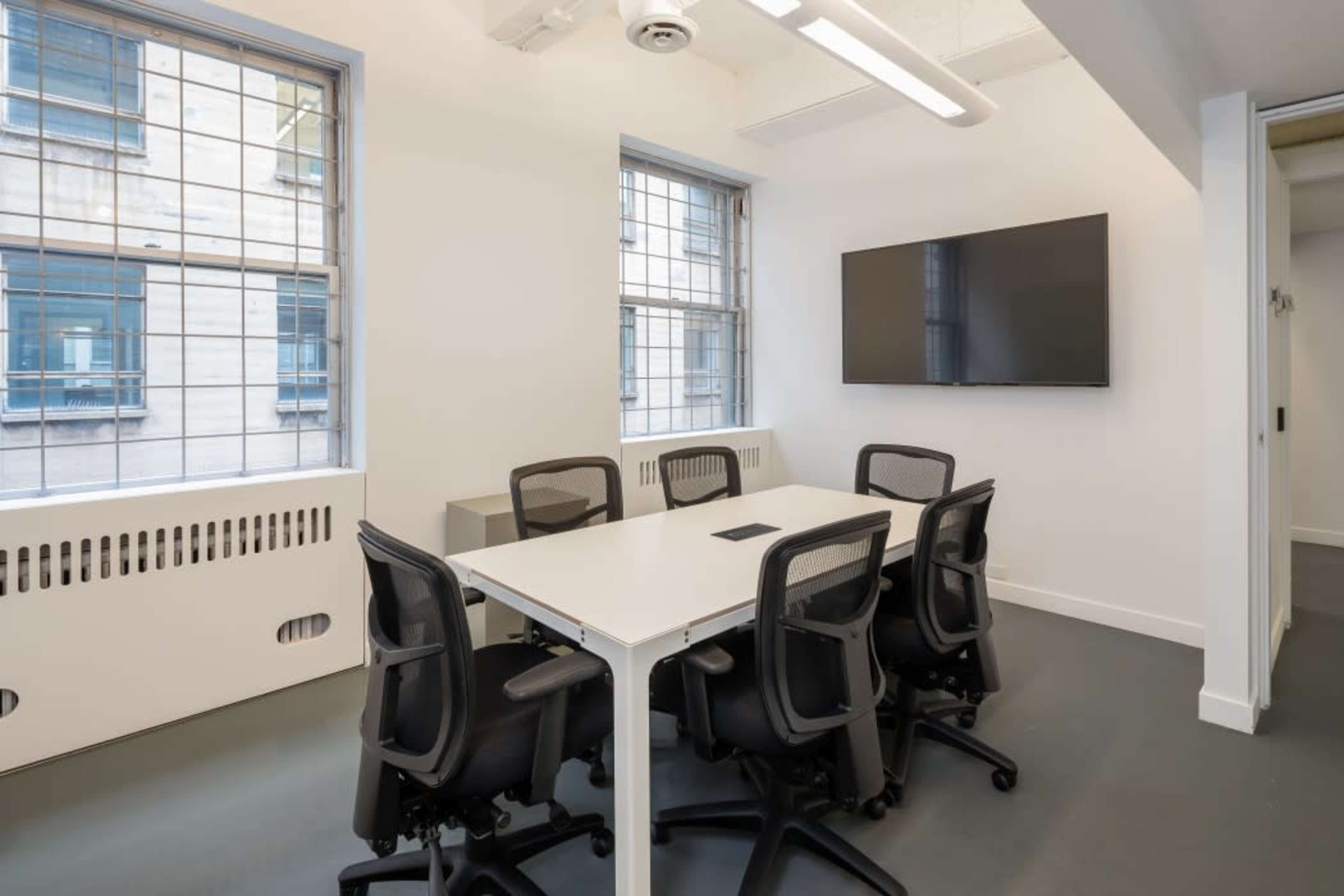 A modern conference room features a white table surrounded by black chairs, with a wall-mounted television and large windows providing natural light.