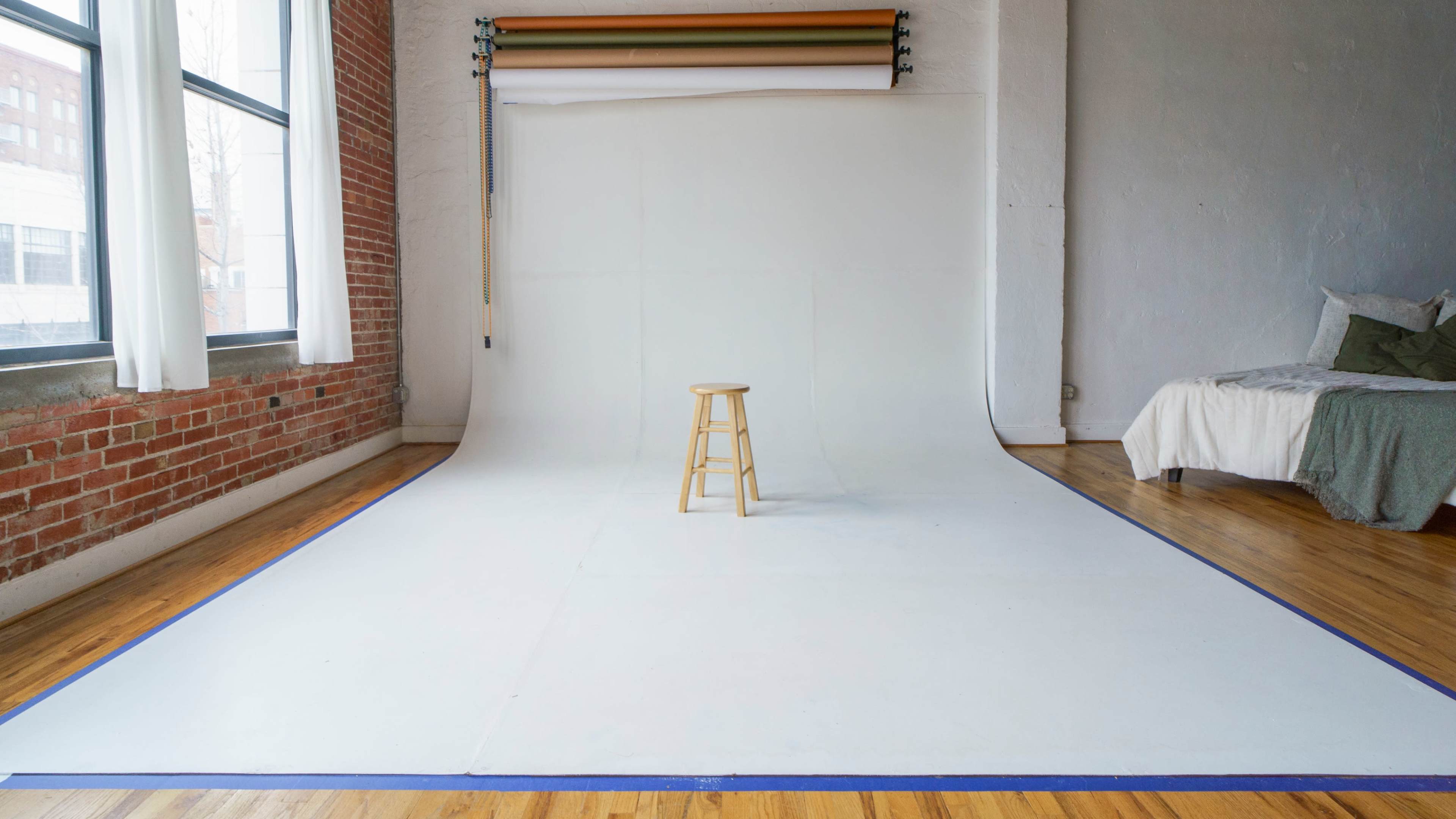 A photography studio with a white backdrop, a wooden stool in the center, and a bed in the background.