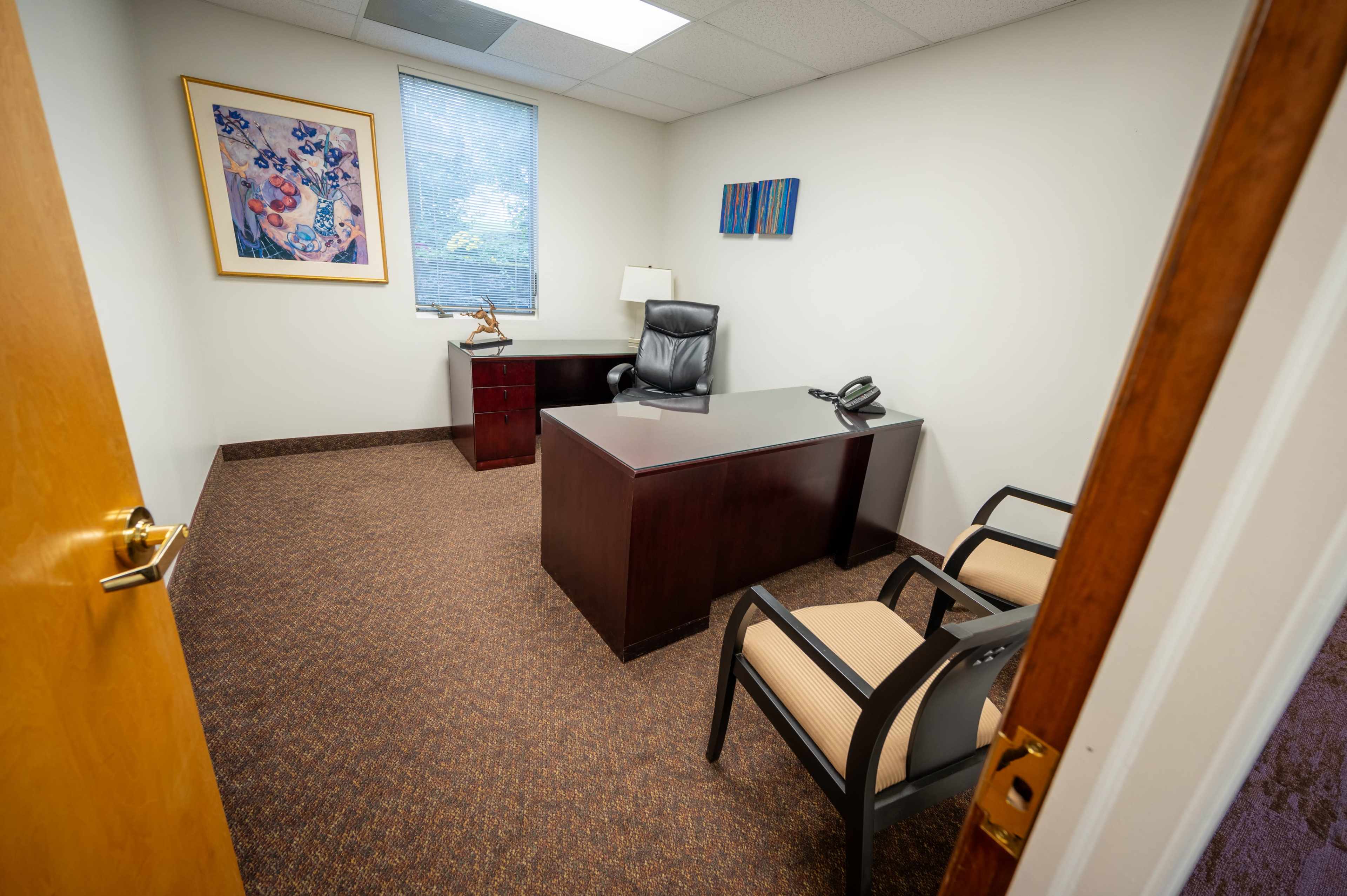 The image shows a small office with a desk, a chair, and two additional chairs, set against a neutral wall and decorated with a piece of artwork.