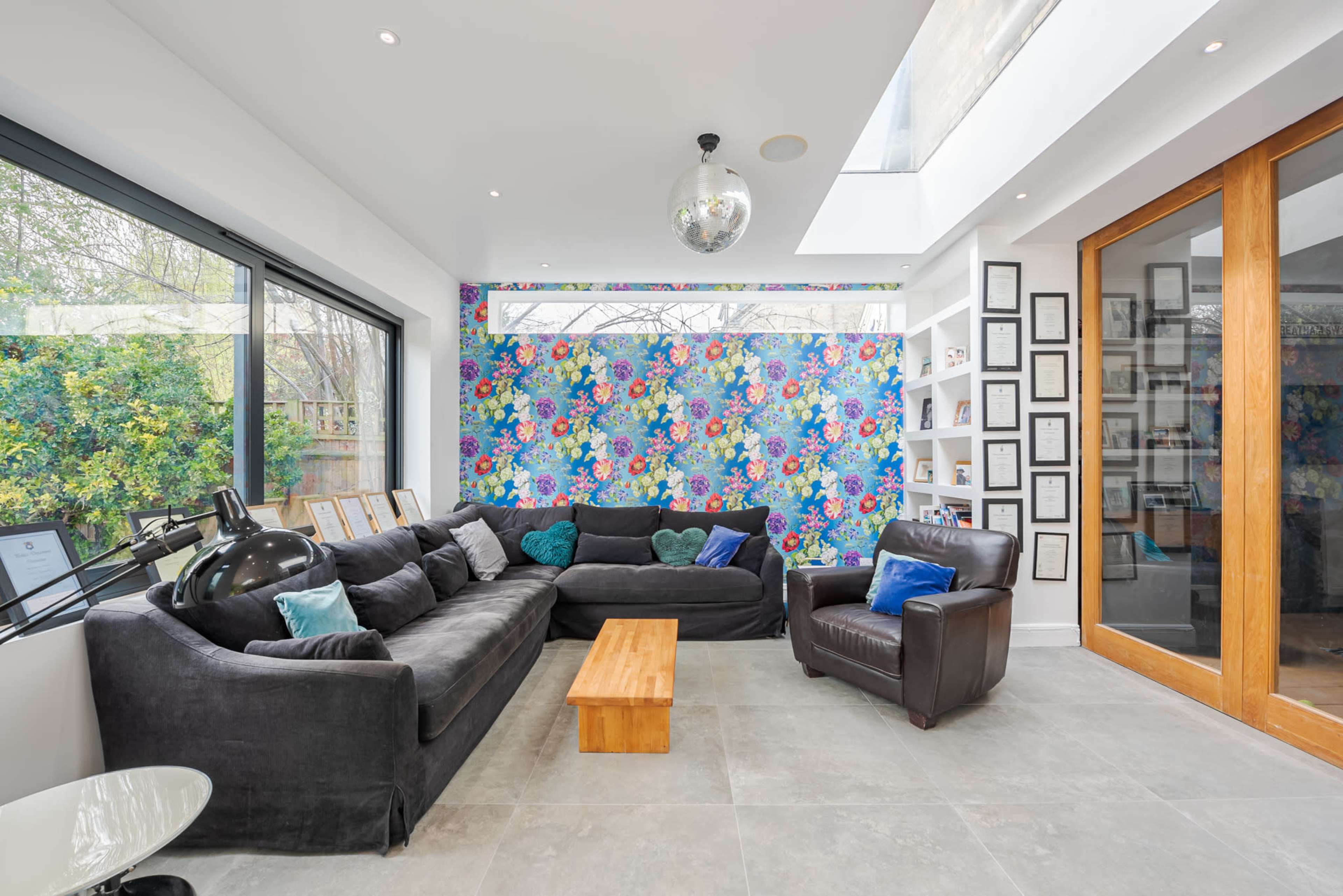 A modern living room features a floral accent wall, large windows, a black sectional sofa, a wooden coffee table, and a disco ball hanging from the ceiling.