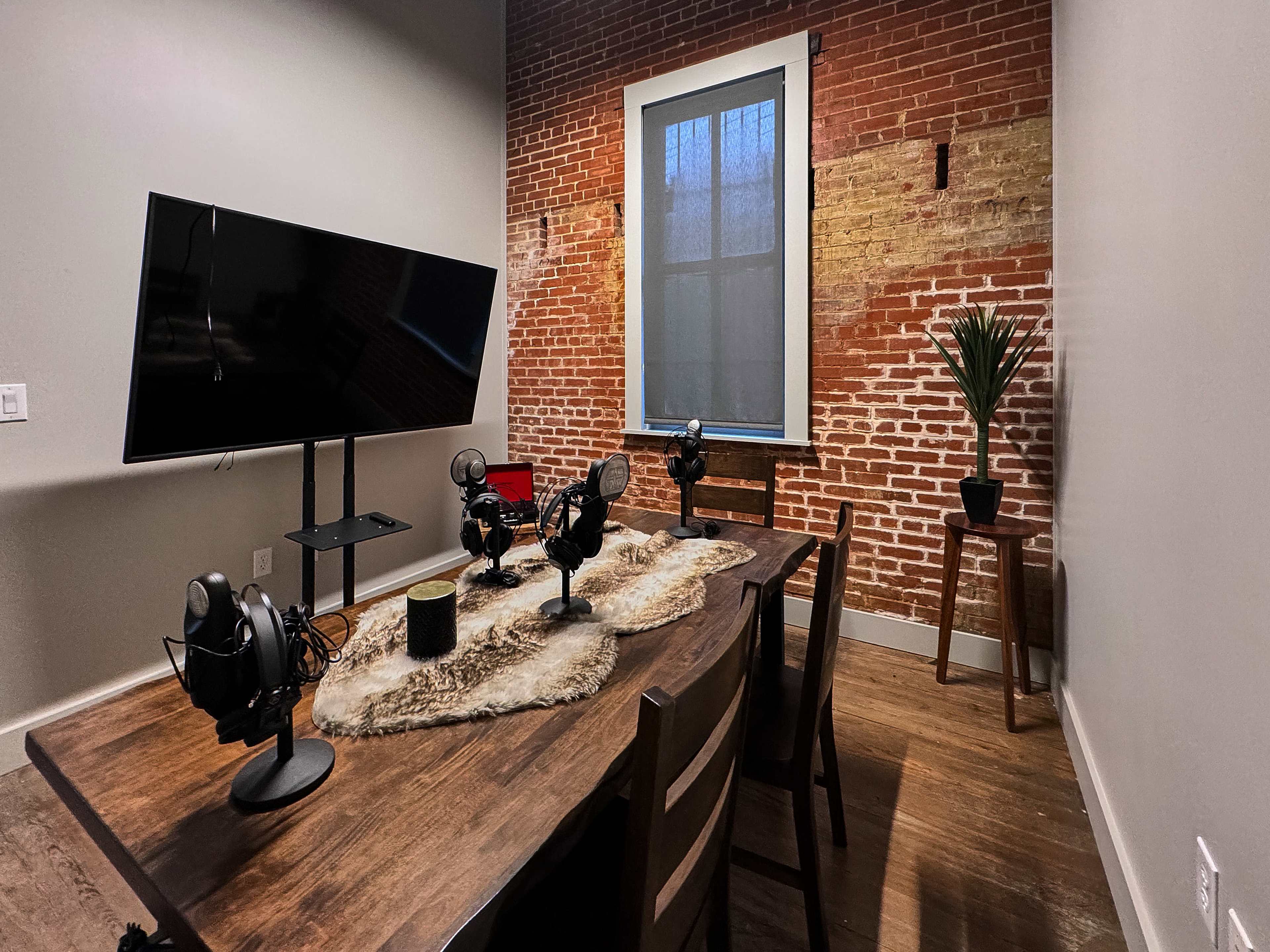 A recording studio features a wooden table with several microphones, a television on a stand, and a potted plant against a brick wall.