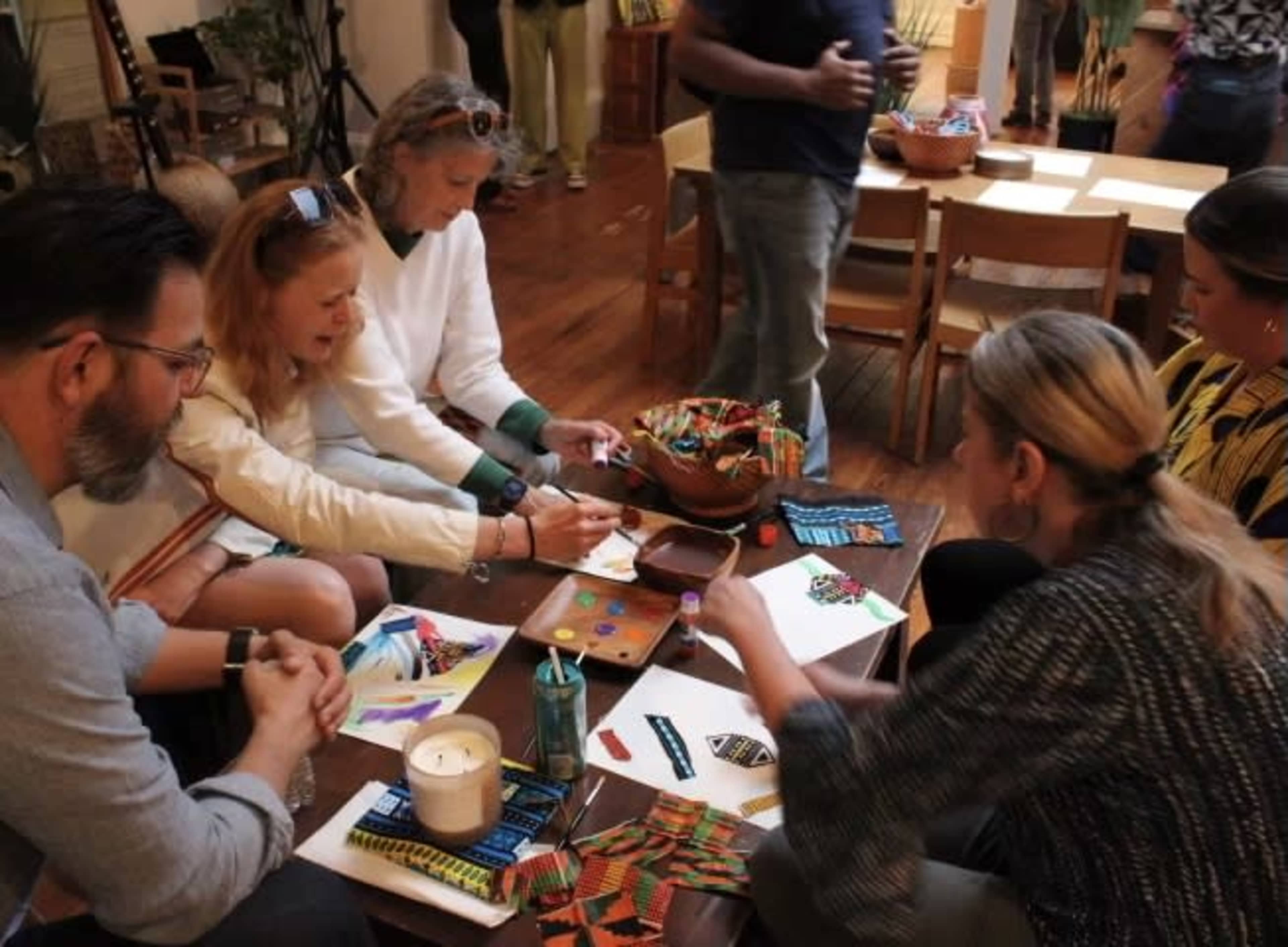 A group of five people is seated around a table engaged in art and craft activities, with various materials and decorated papers spread out before them.