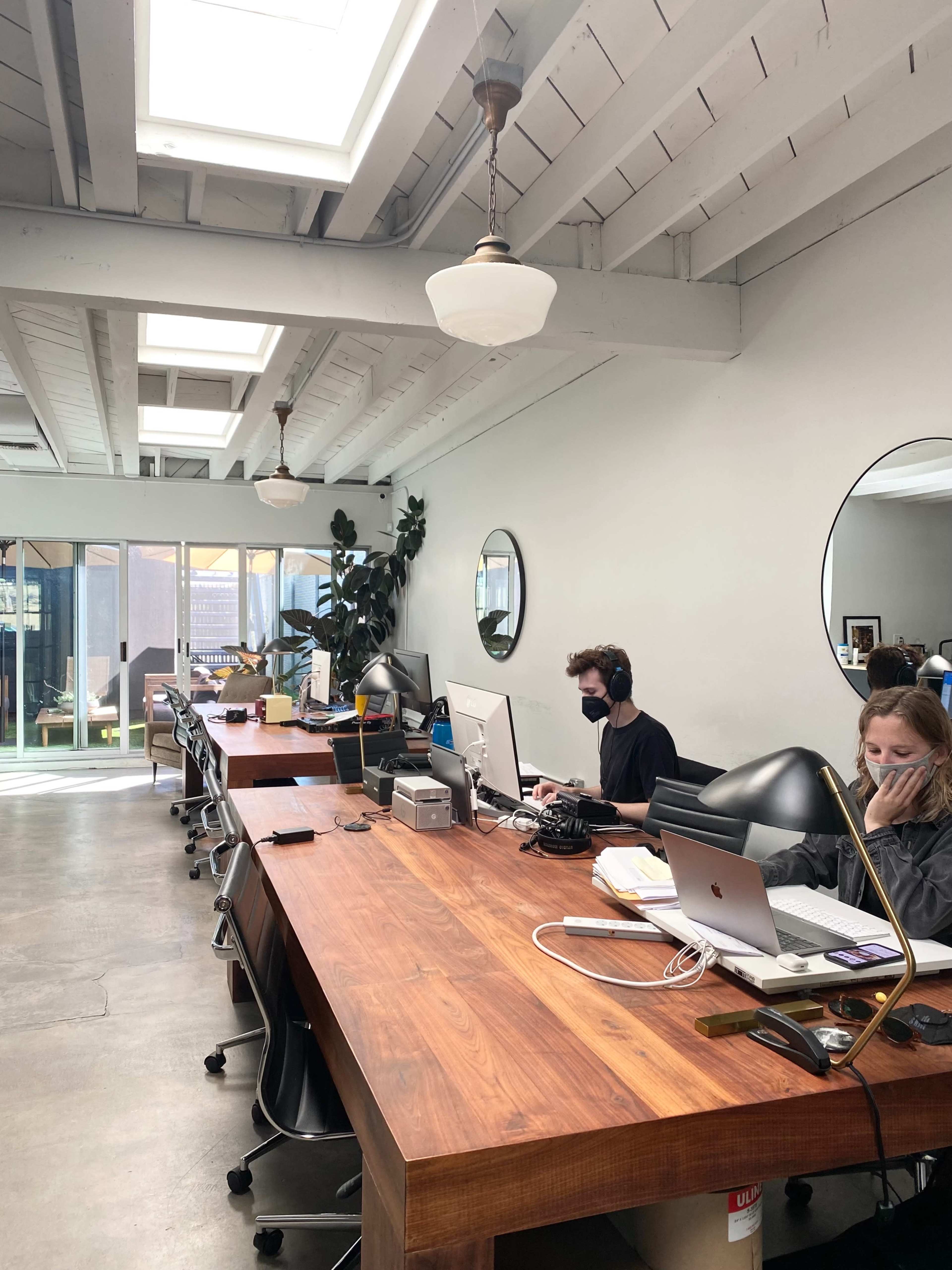 The image shows a bright workspace with a long wooden table, several computers, and two people working while wearing headphones.
