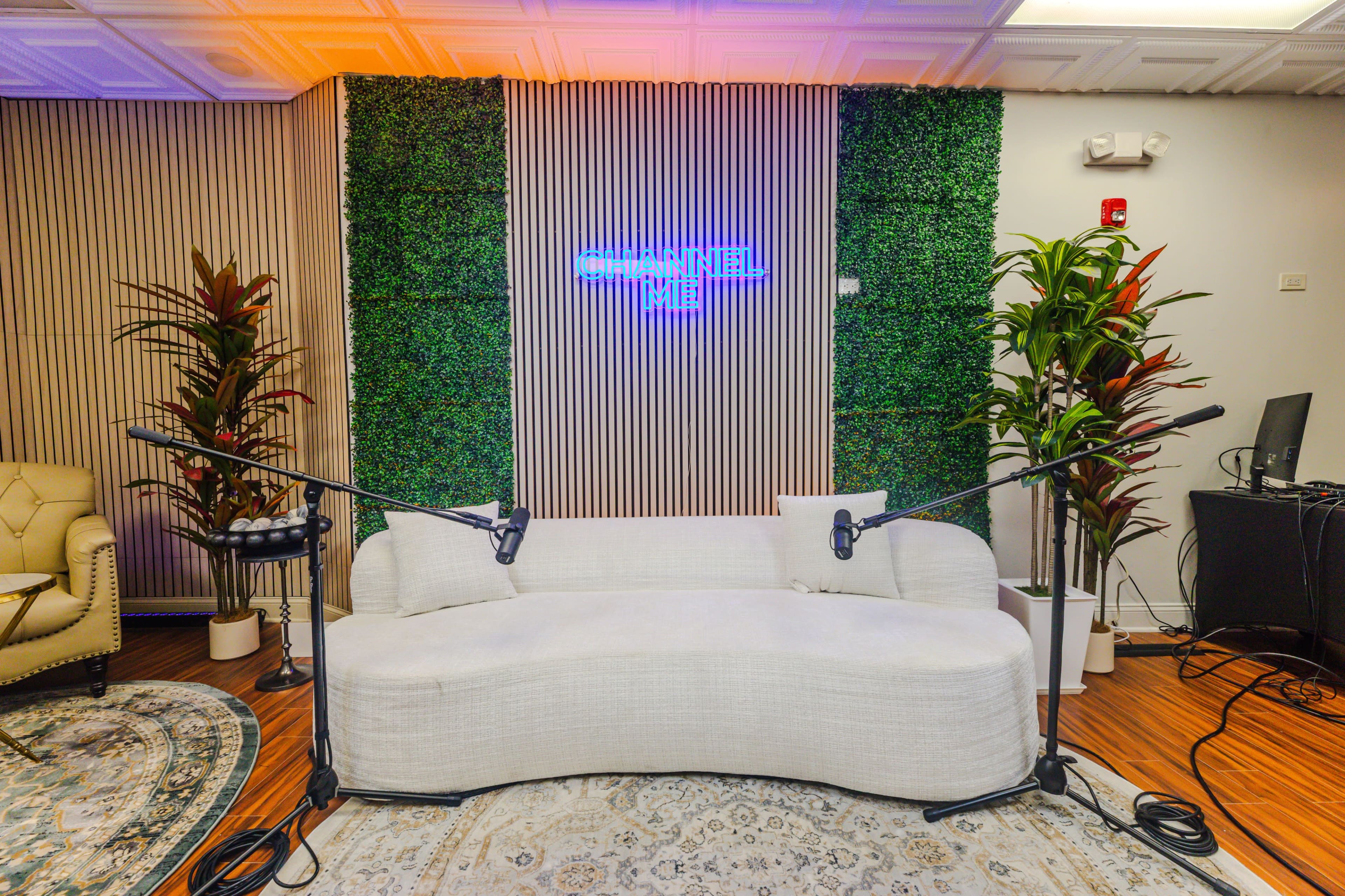 The image shows a modern studio setup with a white curved sofa, two microphones on stands, and green wall foliage, all set against a backdrop featuring a glowing neon sign.
