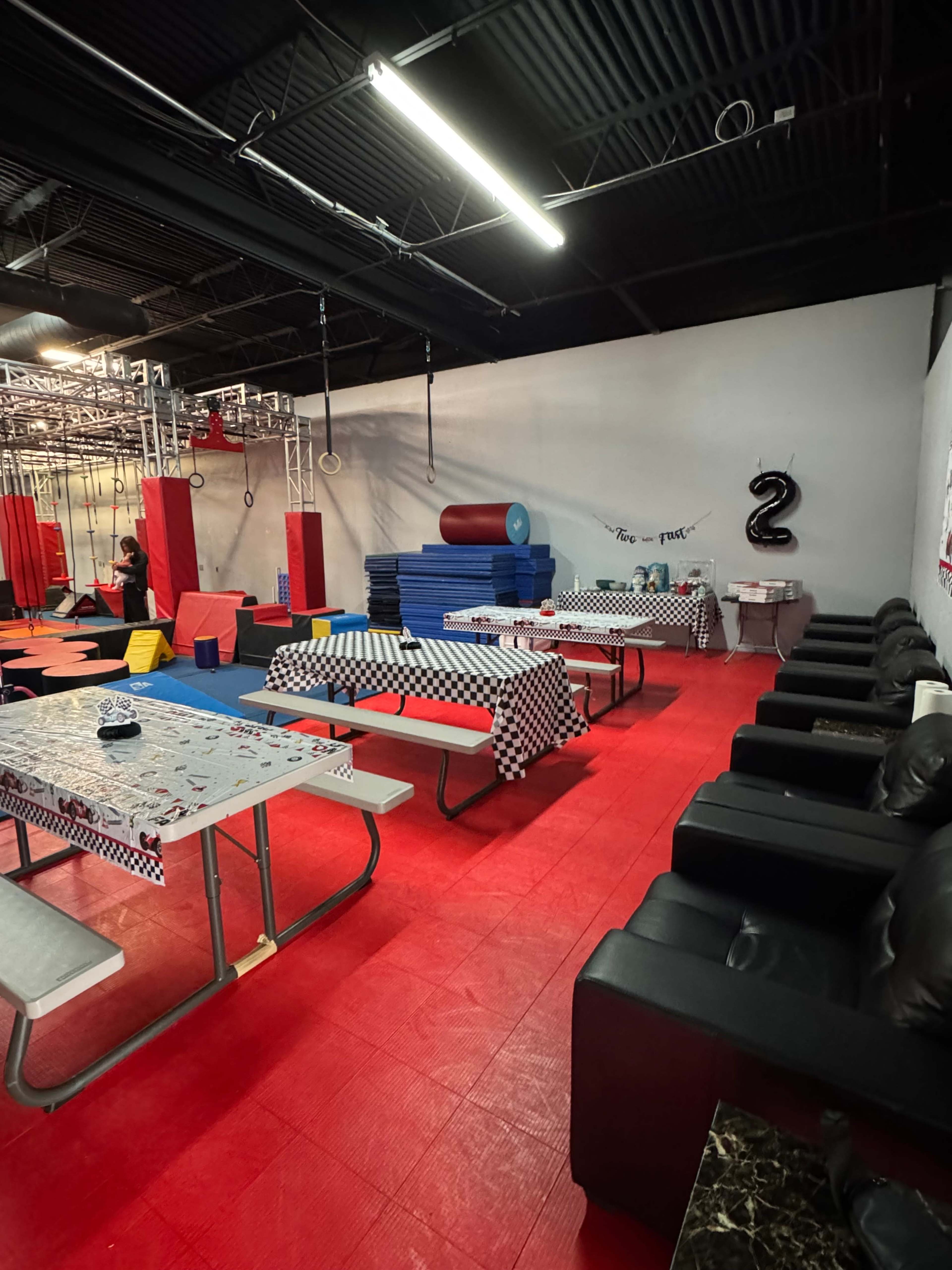 The image shows an indoor space designed for activities with seating areas, tables covered in checkered tablecloths, and various gym equipment in the background.