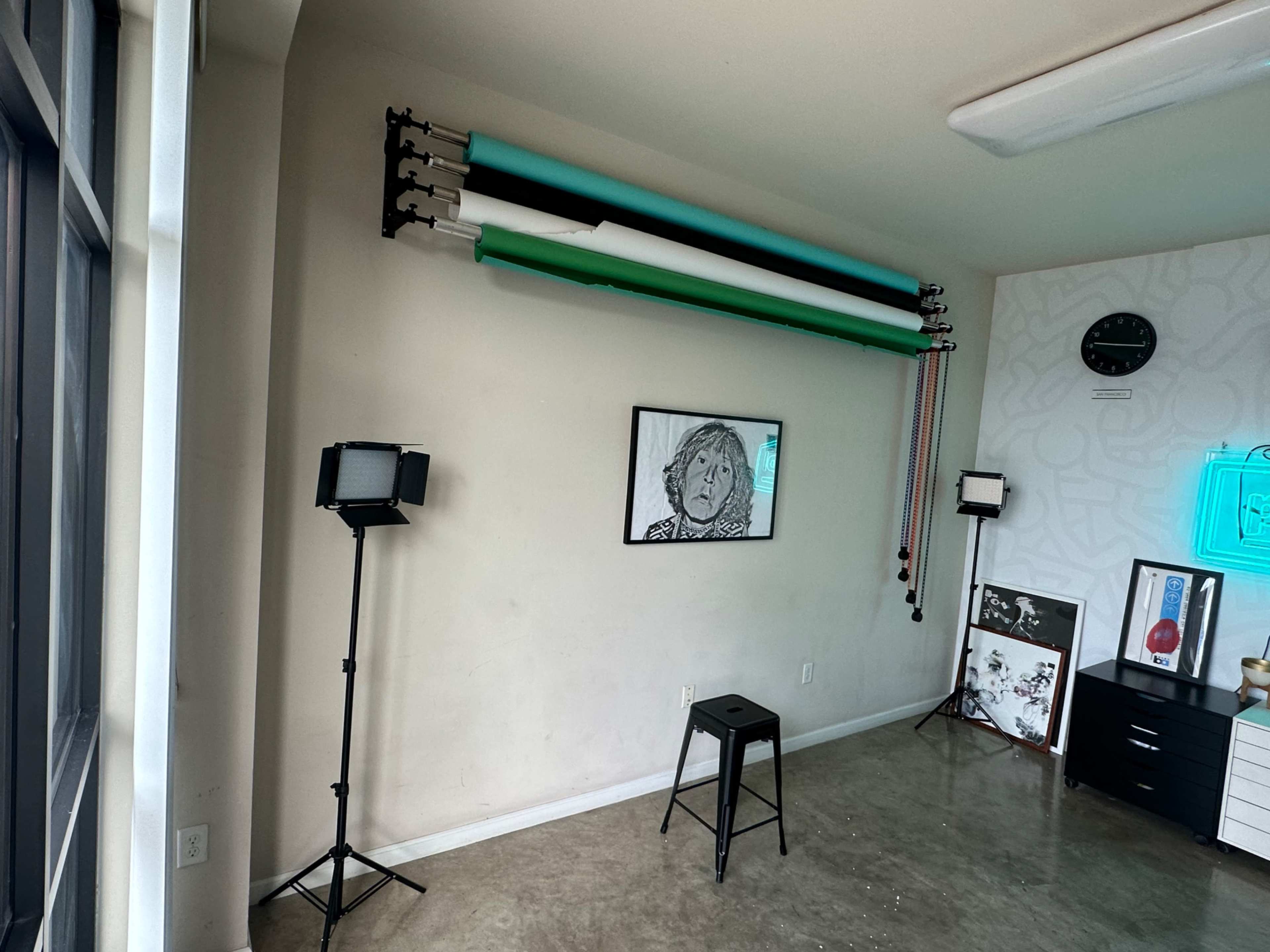A wall with a variety of backdrop rolls mounted above, two studio lights on stands, a stool, and a framed drawing in a room with a modern aesthetic.