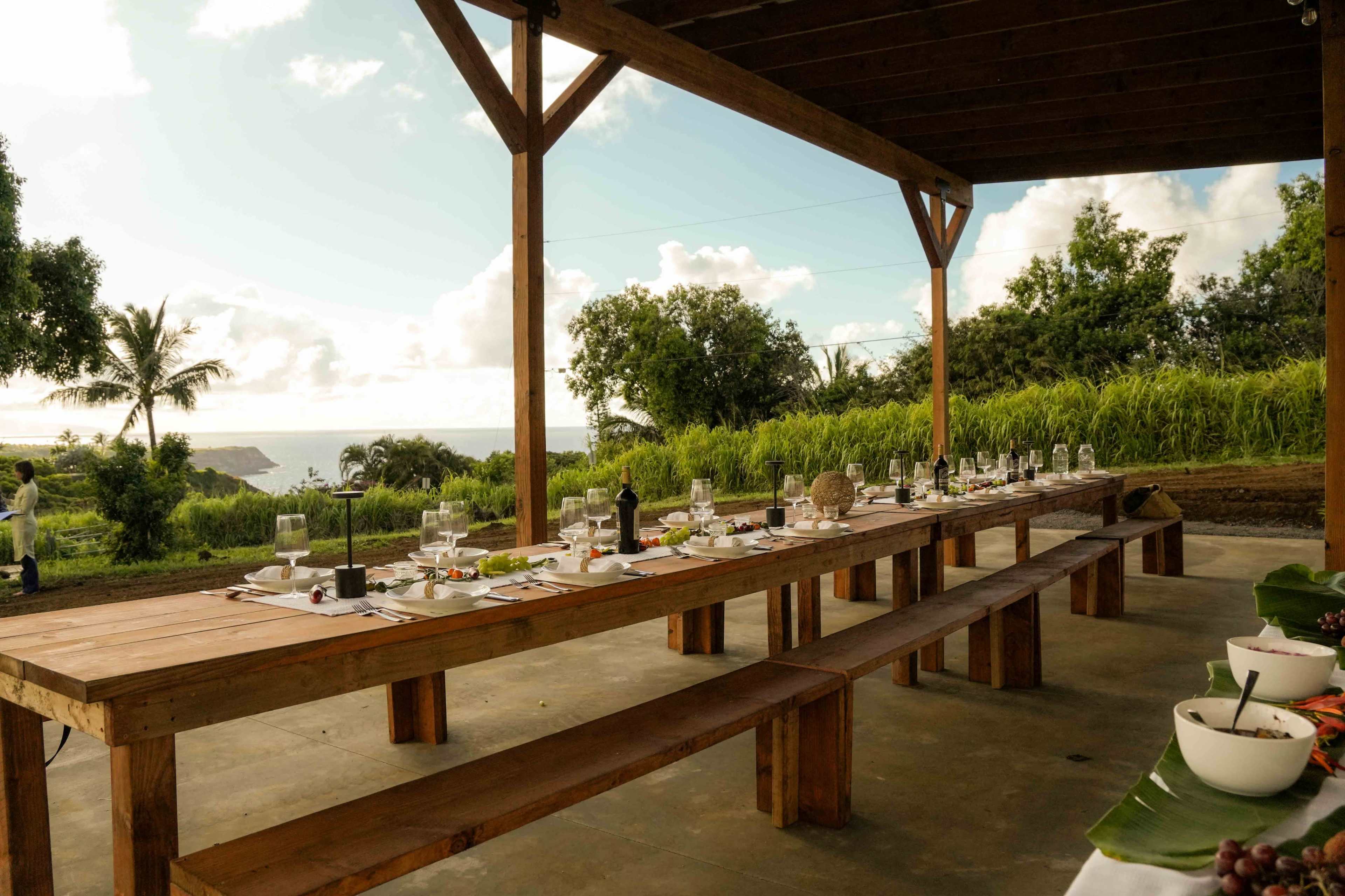 A long wooden table is set for a meal on an outdoor patio overlooking a lush landscape and the ocean in the distance.