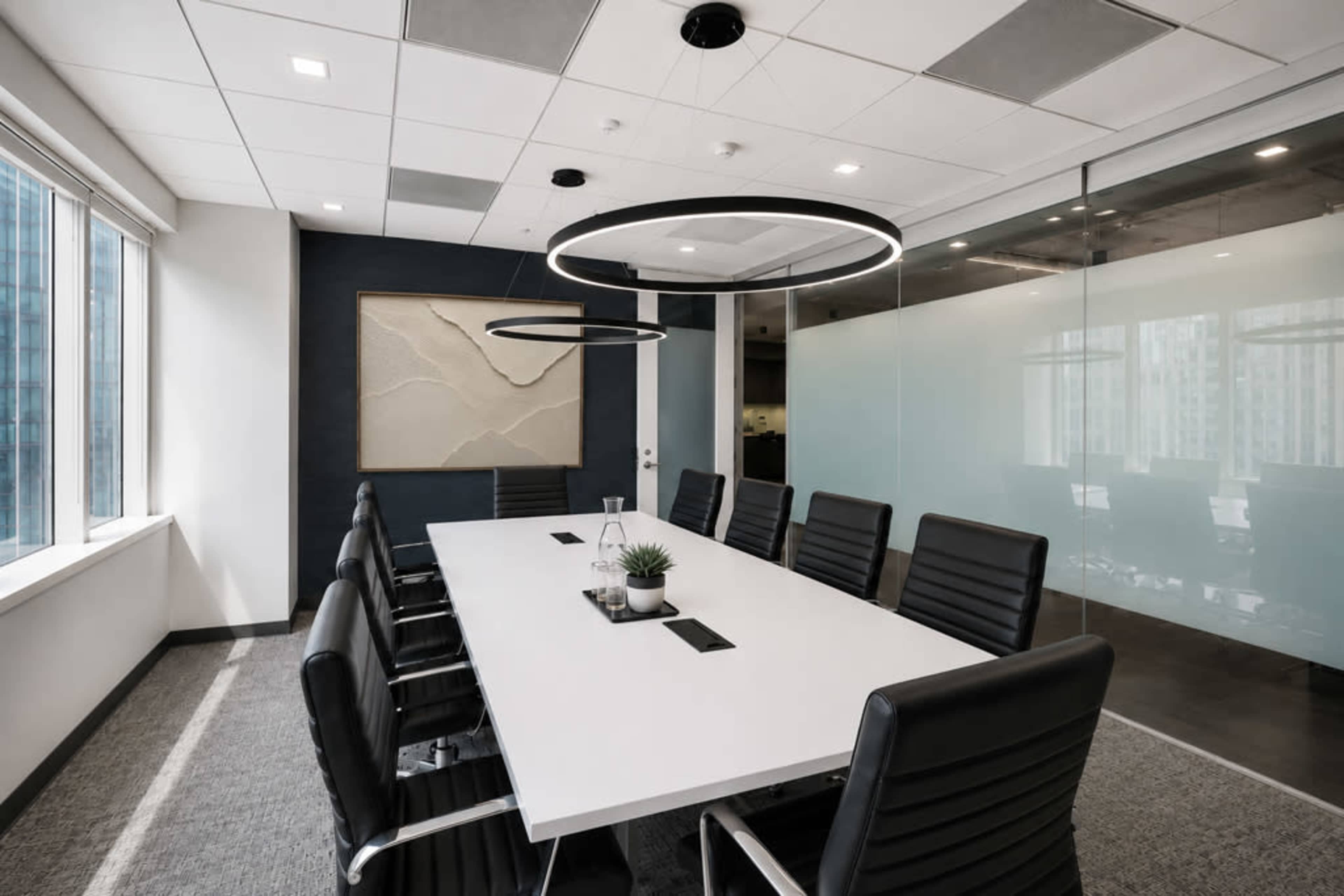 A modern conference room features a large white table surrounded by black leather chairs and illuminated by circular pendant lights.