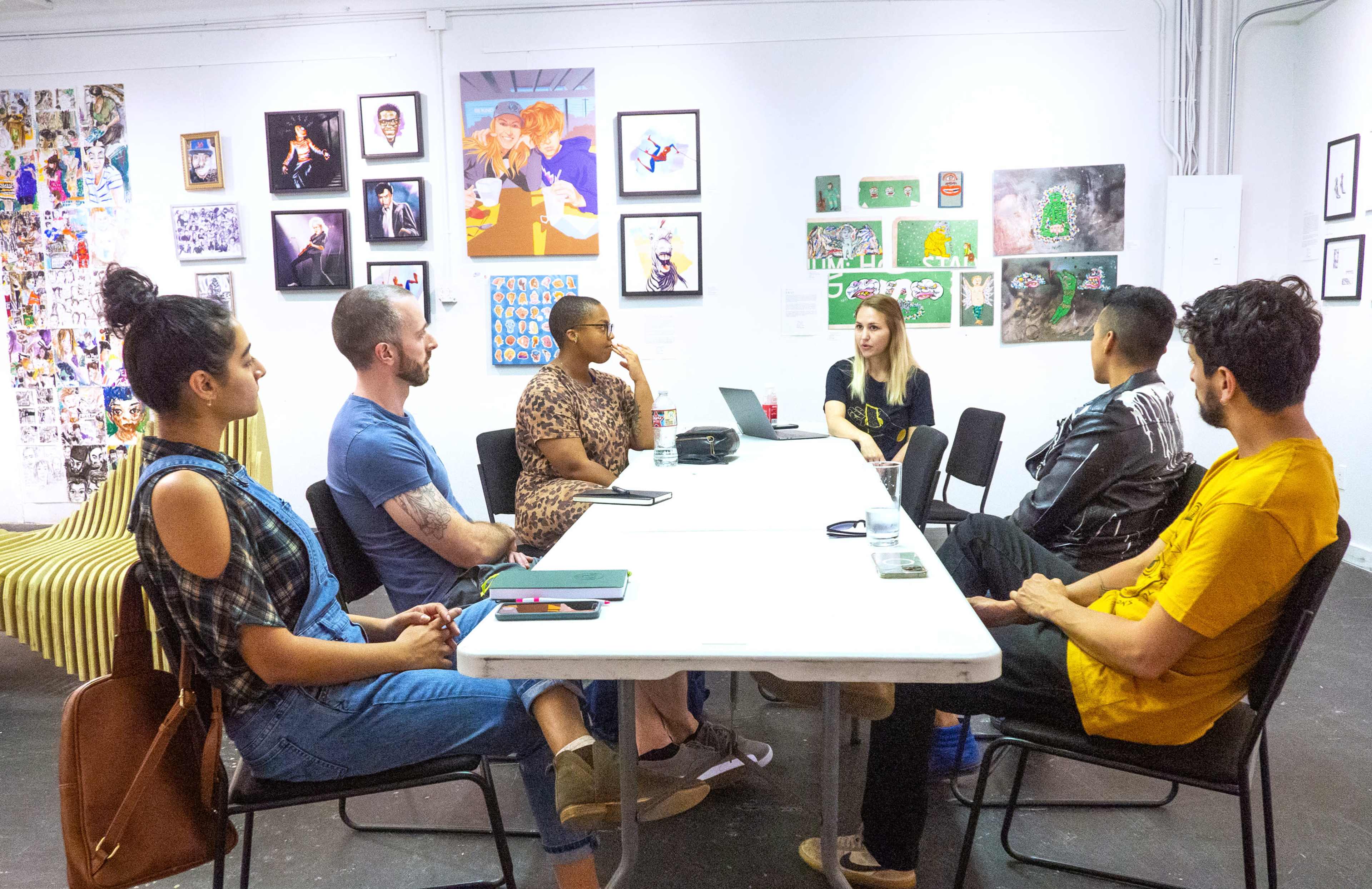 A group of six people is seated around a table in an art gallery setting, engaging in a discussion with various artworks displayed on the walls.